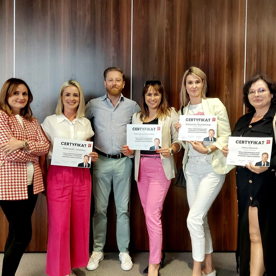 Grupa agentów nieruchomości prezentuje certyfikaty ukończenia kursu, wszyscy uśmiechnięci, na certyfikatach widoczne zdjęcia osób, drewniana ściana w tle.