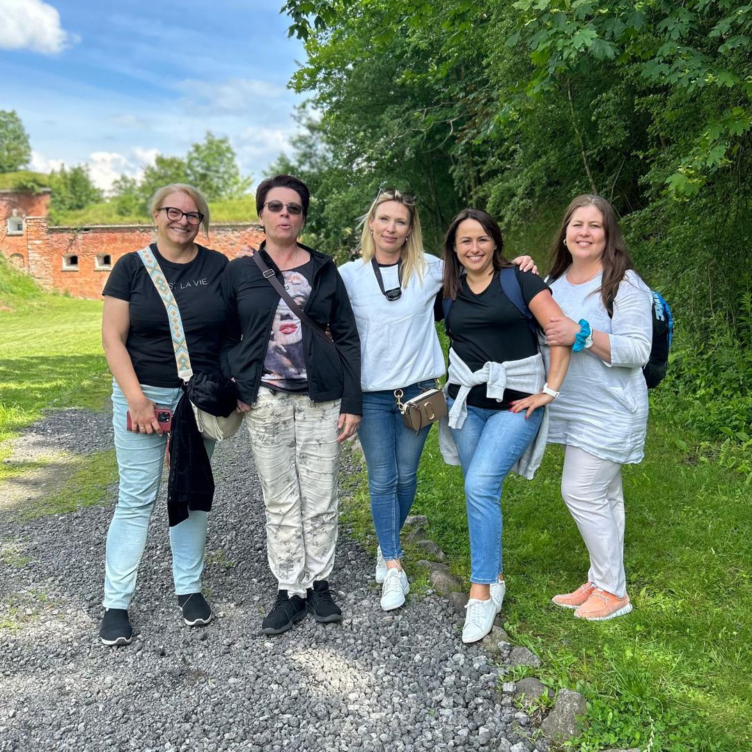 Grupa pięciu kobiet pozuje do zdjęcia na żwirowej ścieżce przed ceglanym budynkiem i drzewami w słoneczny dzień.