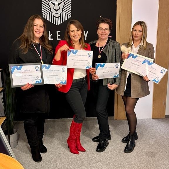 Cztery kobiety prezentują dyplomy uznania na tle ściany z logo firmy, trzy z nich mają na szyjach medale.