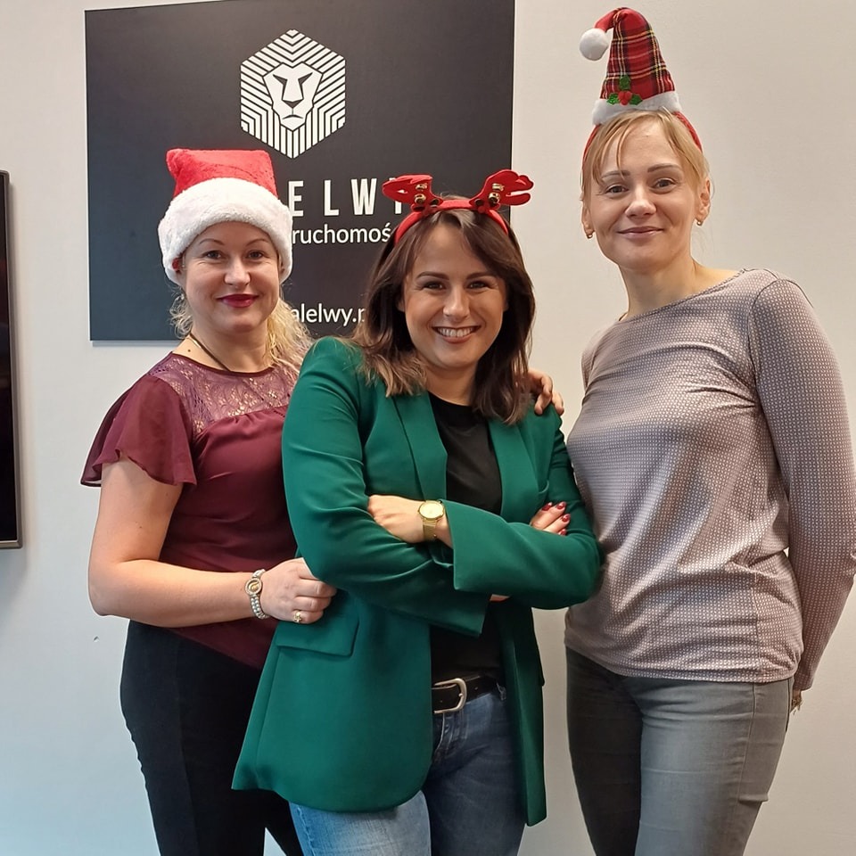 Trzy uśmiechnięte kobiety w świątecznych nakryciach głowy pozują na tle ściany z logo firmy Elelwy Nieruchomości.
