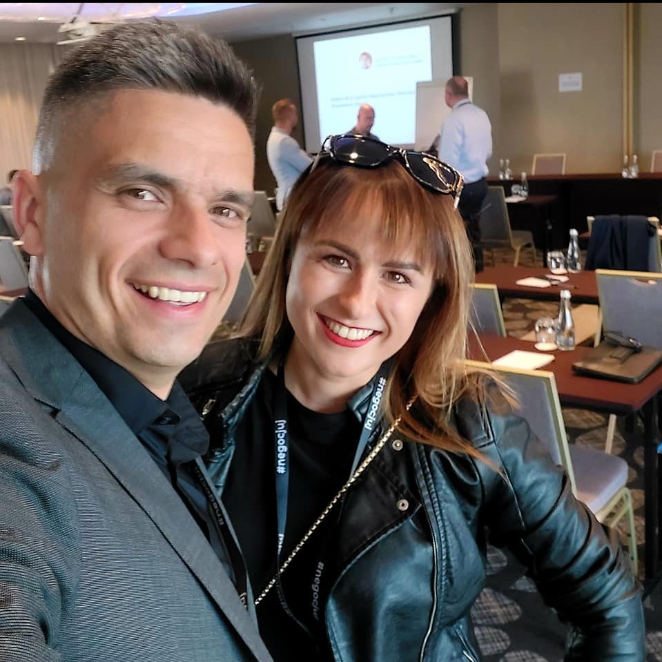 Uśmiechnięci mężczyzna w szarym garniturze i kobieta w czarnej skórzanej kurtce robią selfie na tle sali konferencyjnej z osobami prowadzącymi prezentację na ekranie.