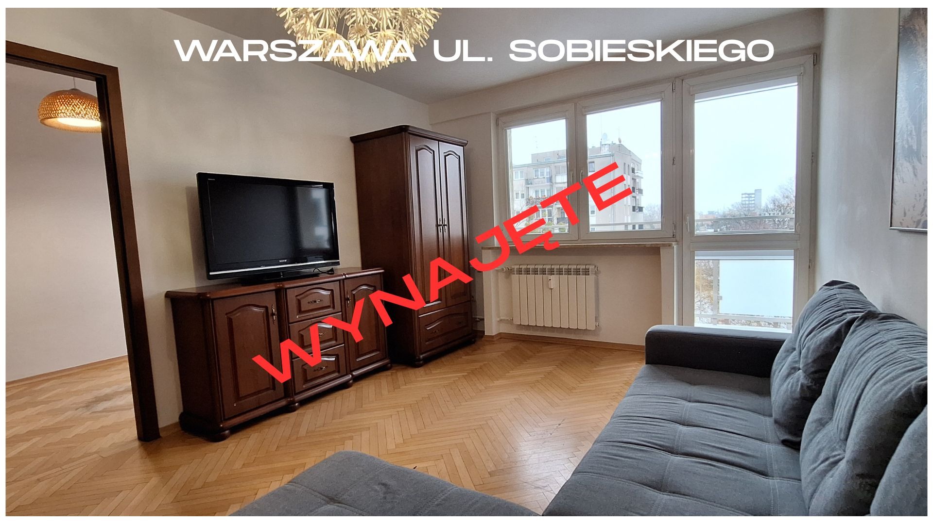 Jasne wnętrze mieszkania na wynajem w Warszawie, z parkietem w jodełkę, meblami w stylu retro i widokiem z okna na miasto. Na zdjęciu widoczny napis 'Wynajęte'.