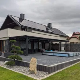 Nowoczesny dom jednorodzinny z panelami fotowoltaicznymi na dachu, zadaszonym tarasem i zadaszonym basenem, otoczony zadbanym ogrodem z ozdobnymi krzewami i kamieniami.