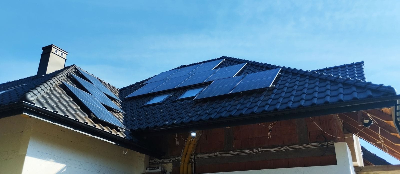 Panele słoneczne zamontowane na dachu domu z czarną dachówką, widoczne okna dachowe i komin na tle błękitnego nieba.