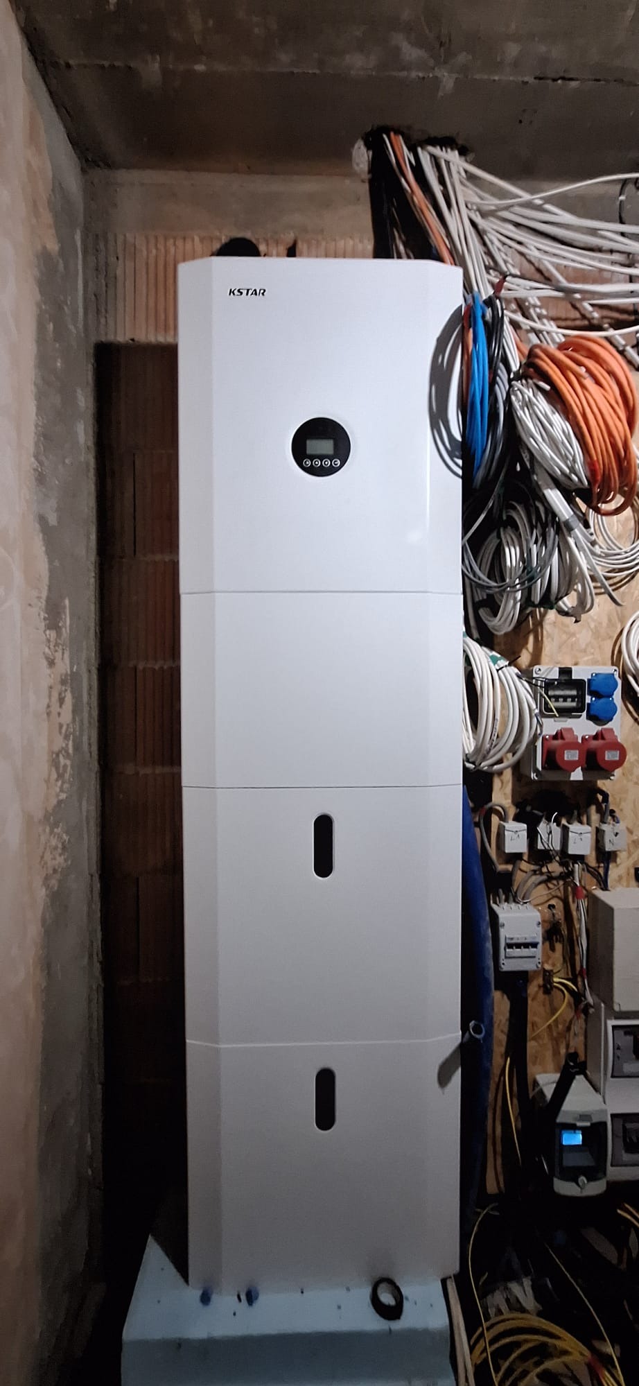 Trzysegmentowy biały magazyn energii KSTAR z wyświetlaczem, zainstalowany w surowym pomieszczeniu z widoczną instalacją elektryczną na ścianie z desek.