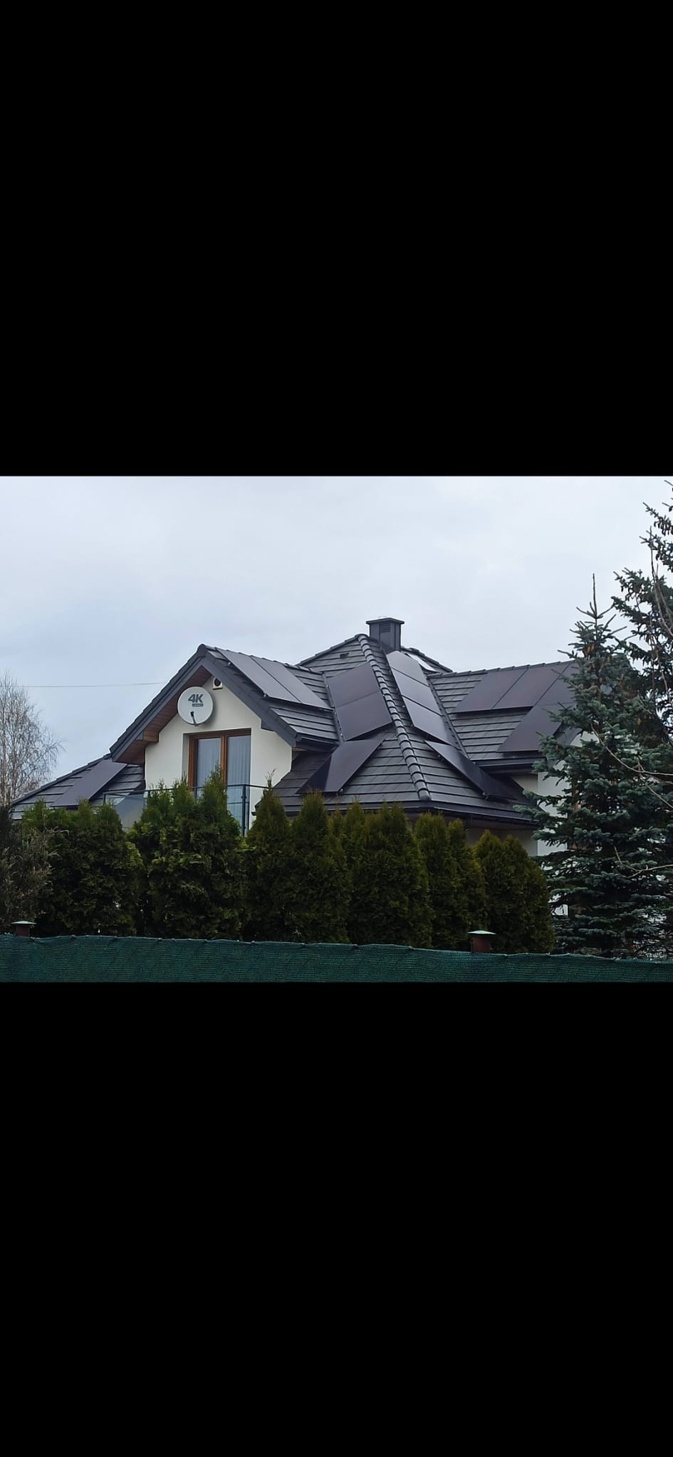 Dom jednorodzinny z ciemną dachówką i panelami na dachu, widoczna antena satelitarna oraz rząd zielonych tuj w ogrodzie.