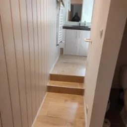 Wnętrze łazienki z drewnopodobnymi płytkami na podłodze i ścianach, widoczne schodki prowadzące do wyższego poziomu, biały grzejnik drabinkowy i fragment umywalki z szafką w lustrze.