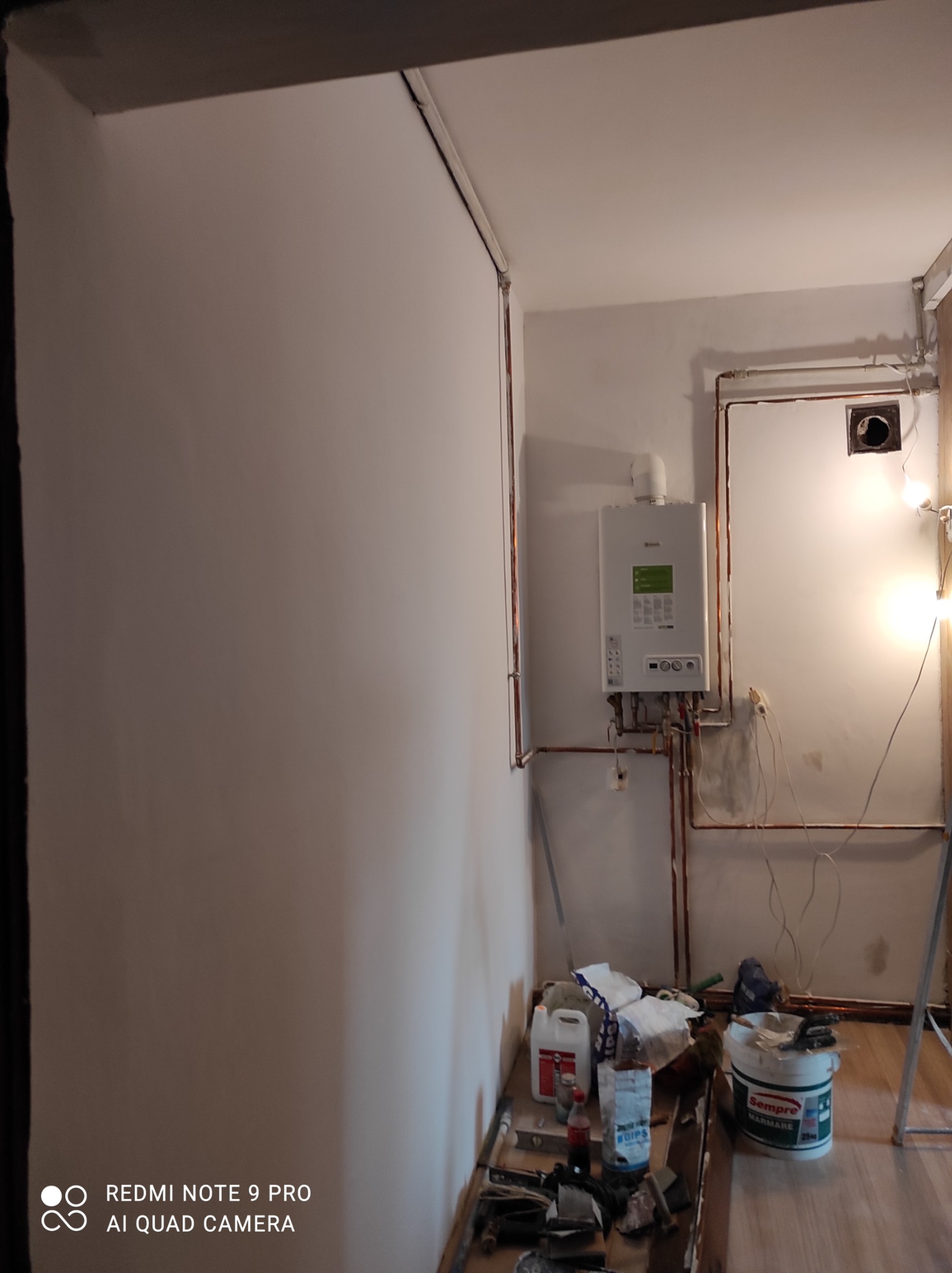 Wnętrze w trakcie prac instalacyjnych: nowy piec gazowy zamontowany na ścianie, widoczne miedziane rury, przewody elektryczne, oraz wiadro z zaprawą i narzędzia na podłodze.