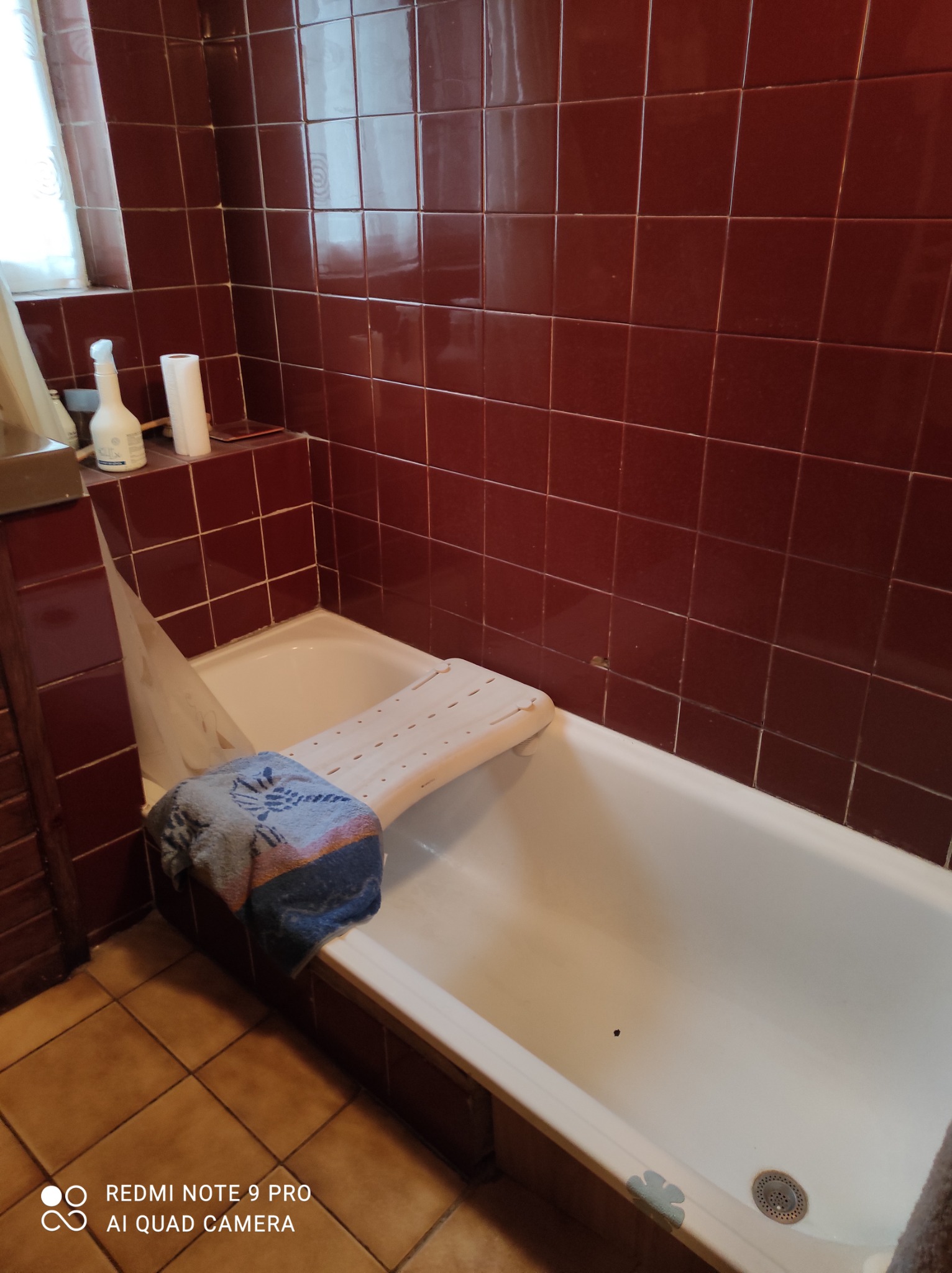 Ciemnoczerwone kafelki w łazience z białą wanną i niebieskim ręcznikiem na krawędzi. Widoczny narożnik z oknem, biała butelka z dozownikiem i rolka papieru toaletowego na półce.