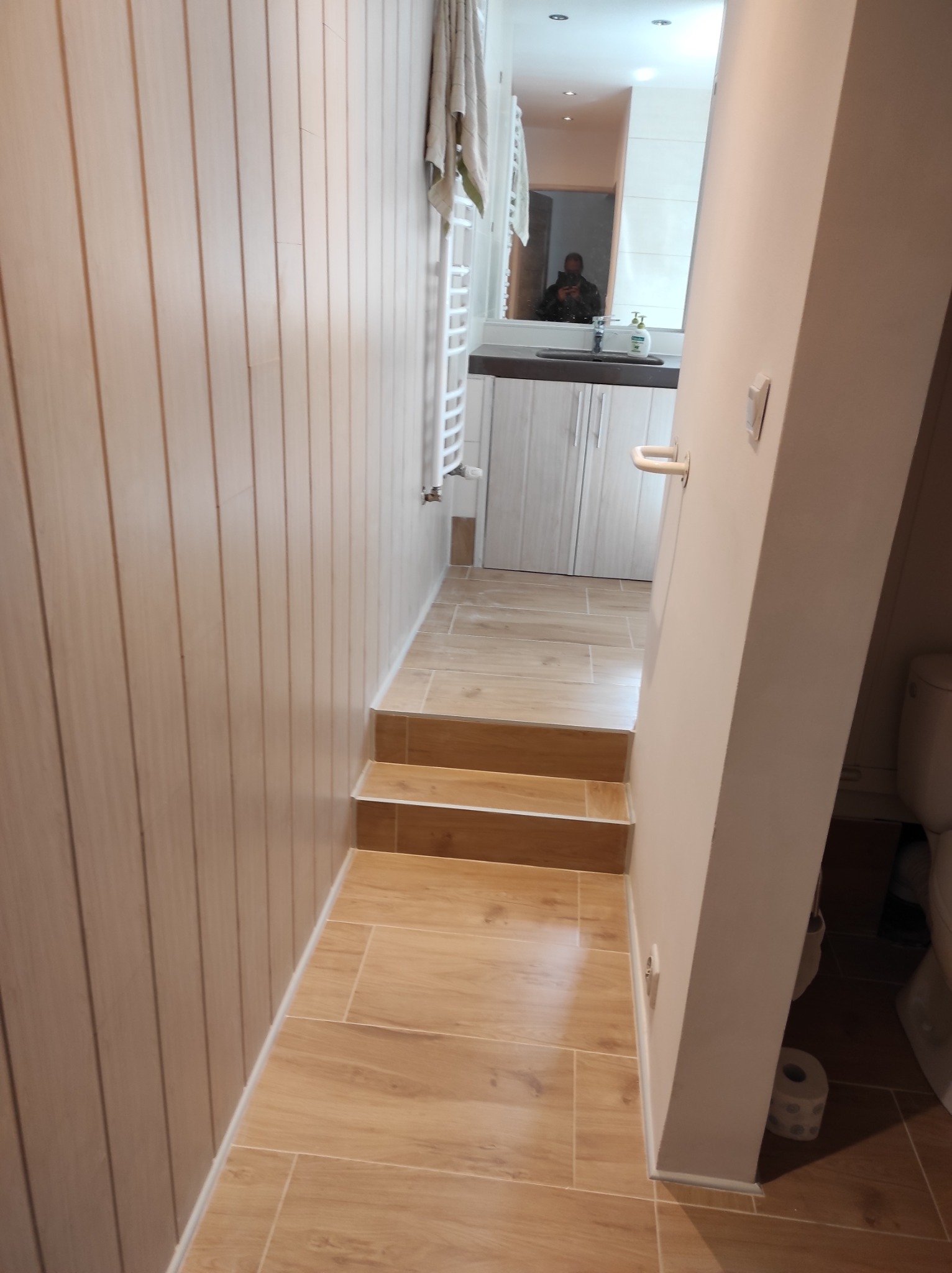 Wnętrze łazienki z drewnopodobnymi płytkami na podłodze i ścianach, widoczne schodki prowadzące do wyższego poziomu, biały grzejnik drabinkowy i fragment umywalki z szafką w lustrze.