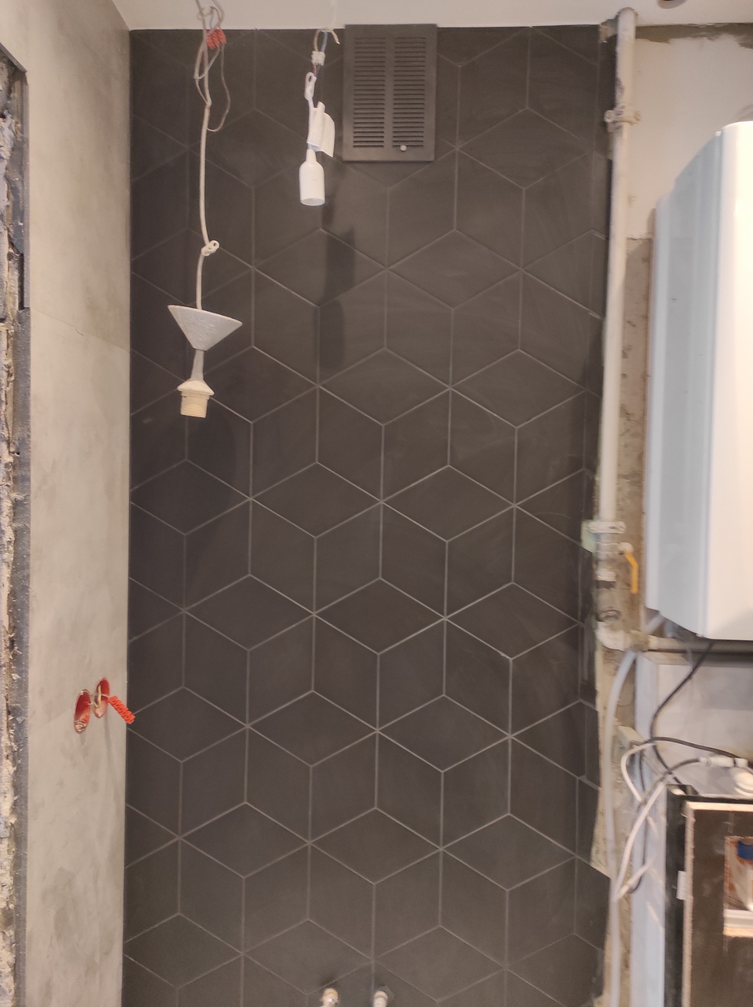Ciemne płytki heksagonalne na ścianie łazienki w trakcie remontu, widoczne kable elektryczne, grzejnik i surowy beton na ścianach.