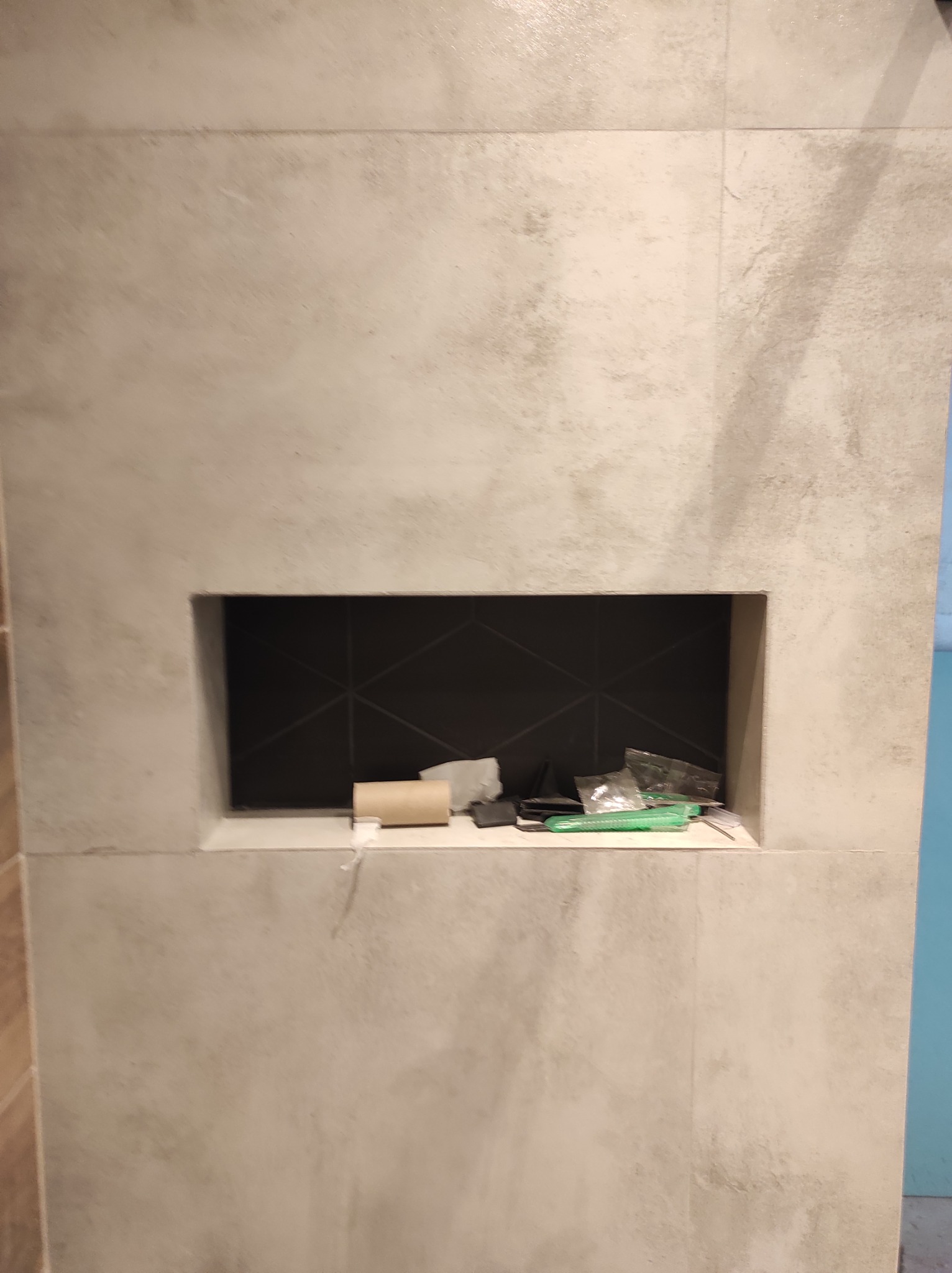 Niewielka wnęka wyłożona czarnymi płytkami z geometrycznym wzorem, otoczona beżowymi płytkami imitującymi beton, z widocznymi akcesoriami i odpadami budowlanymi wewnątrz.
