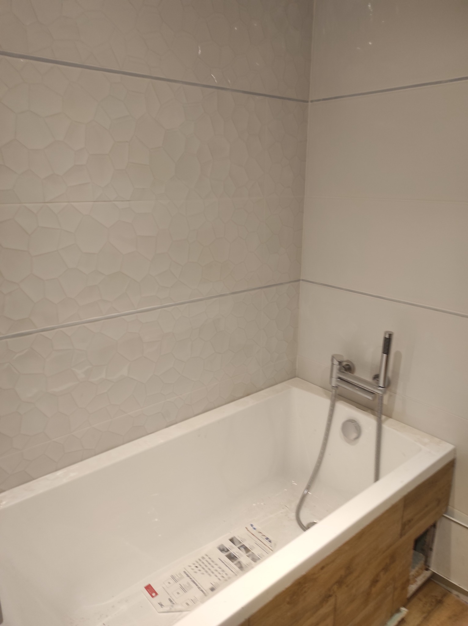 Biała wanna obudowana panelami imitującymi drewno, z baterią i słuchawką prysznicową, otoczona jasnymi płytkami z delikatną fakturą.