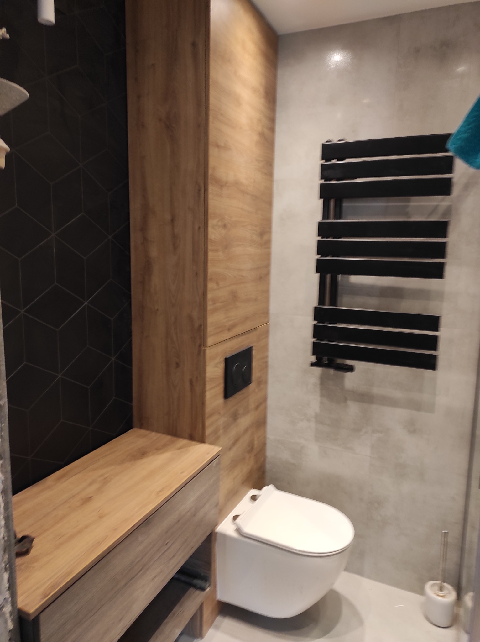 Nowoczesna łazienka z drewnianą zabudową meblową, czarną ścianą z geometrycznym wzorem, białym wiszącym WC i czarnym grzejnikiem drabinkowym na szarej płytce.