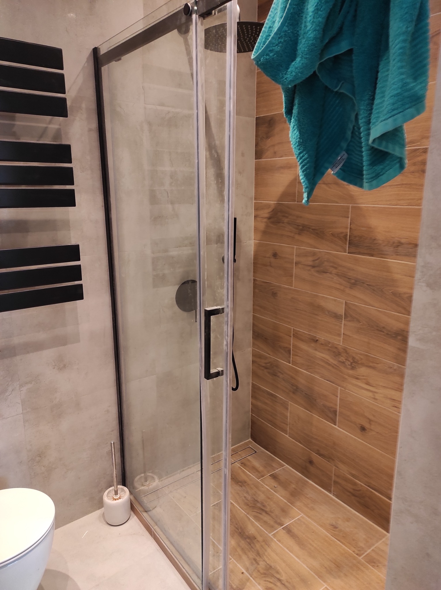 Nowoczesna łazienka z kabiną prysznicową typu walk-in z czarnymi profilami, drewnopodobne płytki na ścianach i podłodze, turkusowy ręcznik, grzejnik drabinkowy w kolorze czarnym i toaleta.