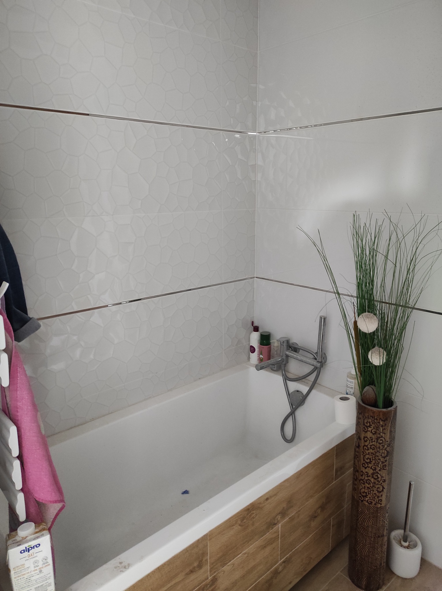 Ukończona łazienka z wanną obudowaną panelami imitującymi drewno, białe płytki z delikatnym wzorem, dekoracyjna trawa w wysokim wazonie i grzejnik drabinkowy z różowym ręcznikiem.