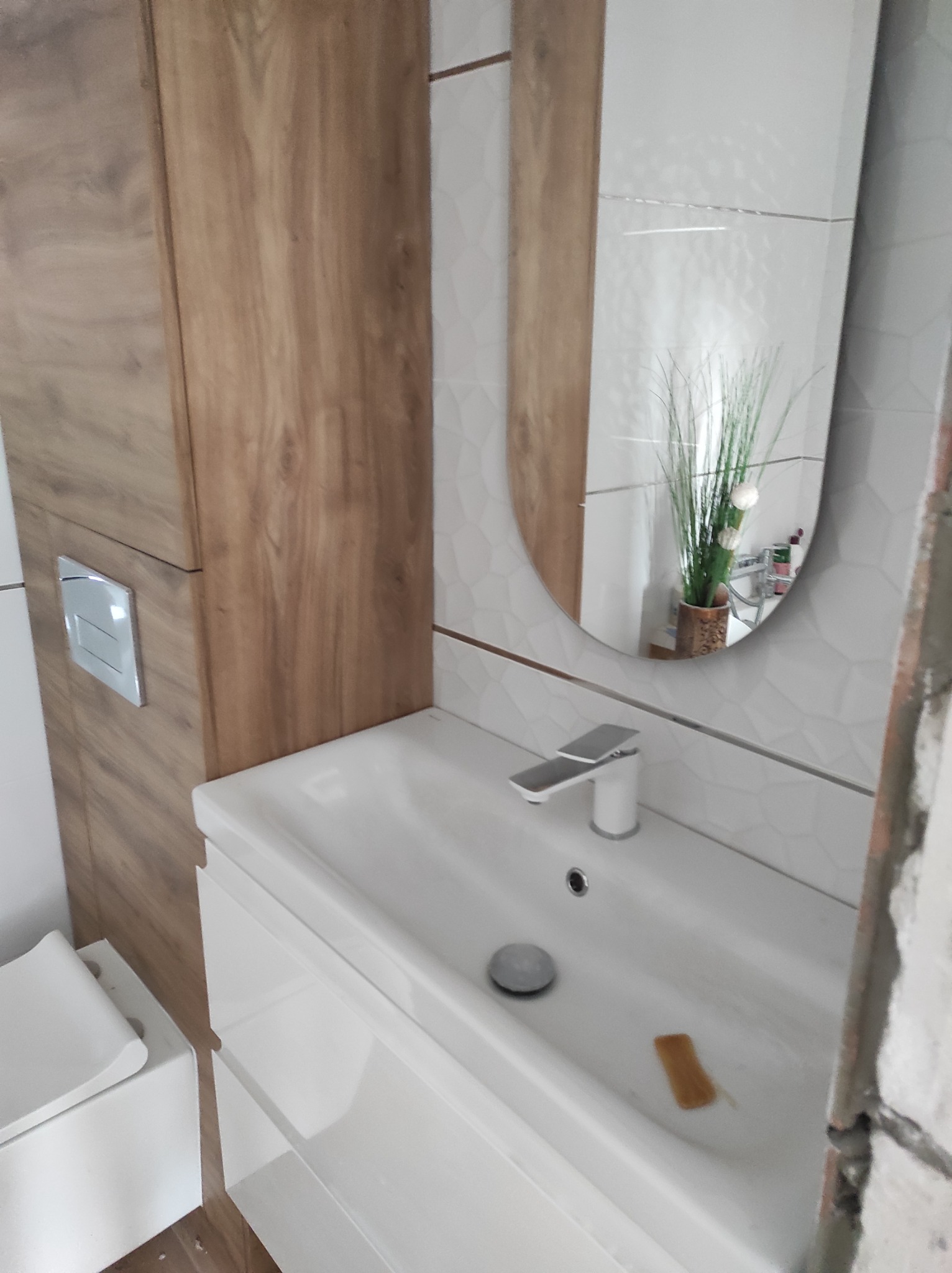 Nowoczesna łazienka w trakcie wykańczania: biała umywalka z szafką, drewnopodobna szafka wisząca, okrągłe lustro odbijające roślinę w doniczce, fragment toalety i surowe wykończenie ściany.