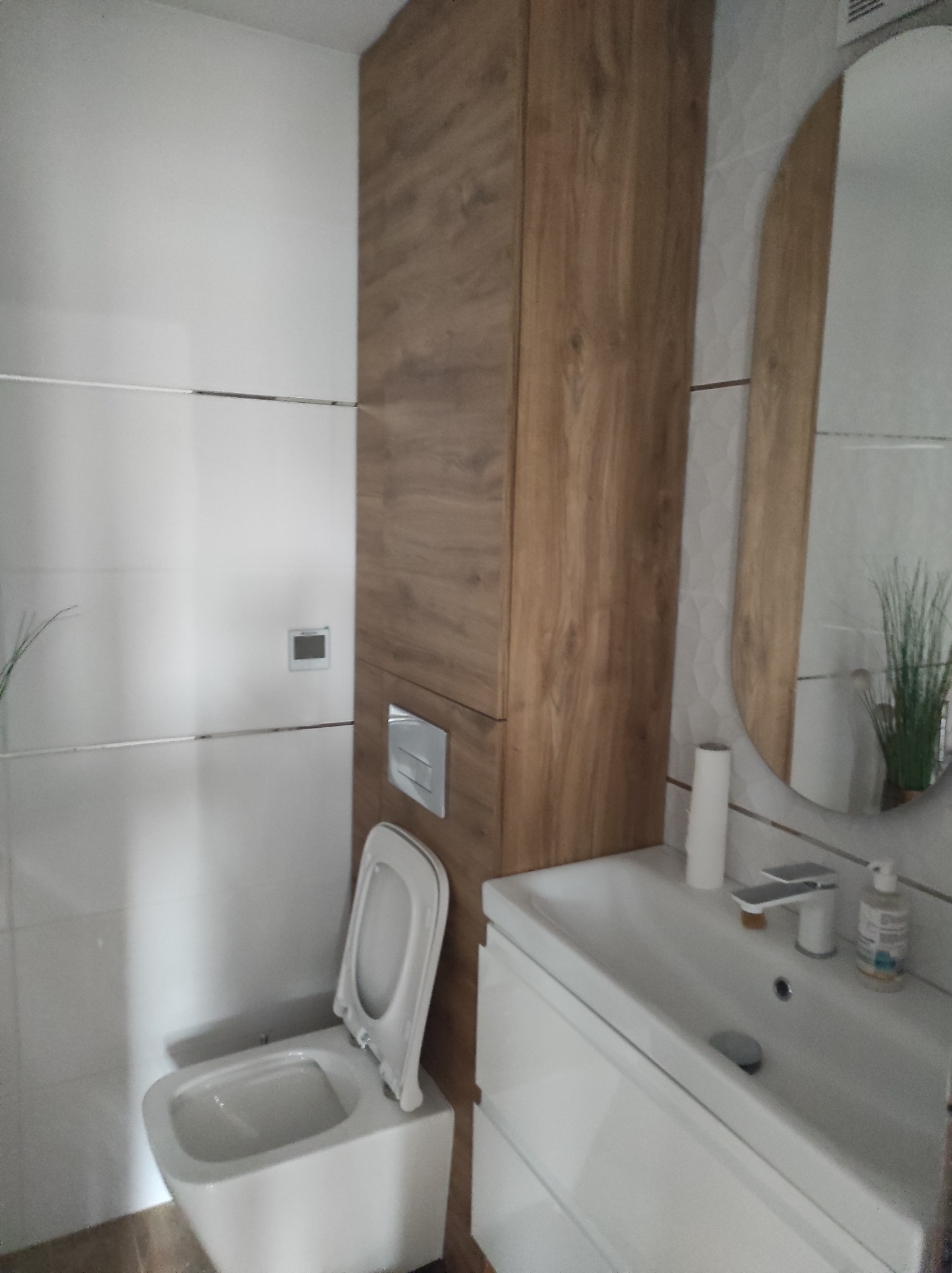 Nowoczesna łazienka z białymi płytkami, podwieszaną toaletą z otwartą klapą, szafką z umywalką i lustrem w drewnianej obudowie, widoczne detale wykończenia.