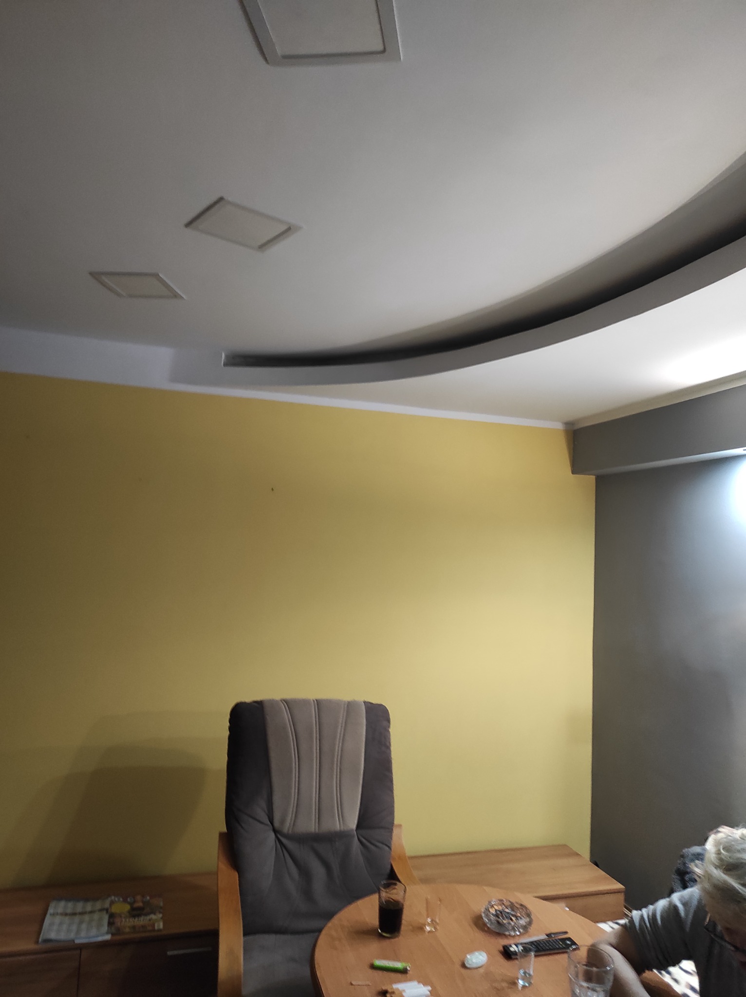 Wnętrze pokoju w Bytomiu z żółtą ścianą, fotelem i fragmentem sufitu z podwieszanym elementem dekoracyjnym.