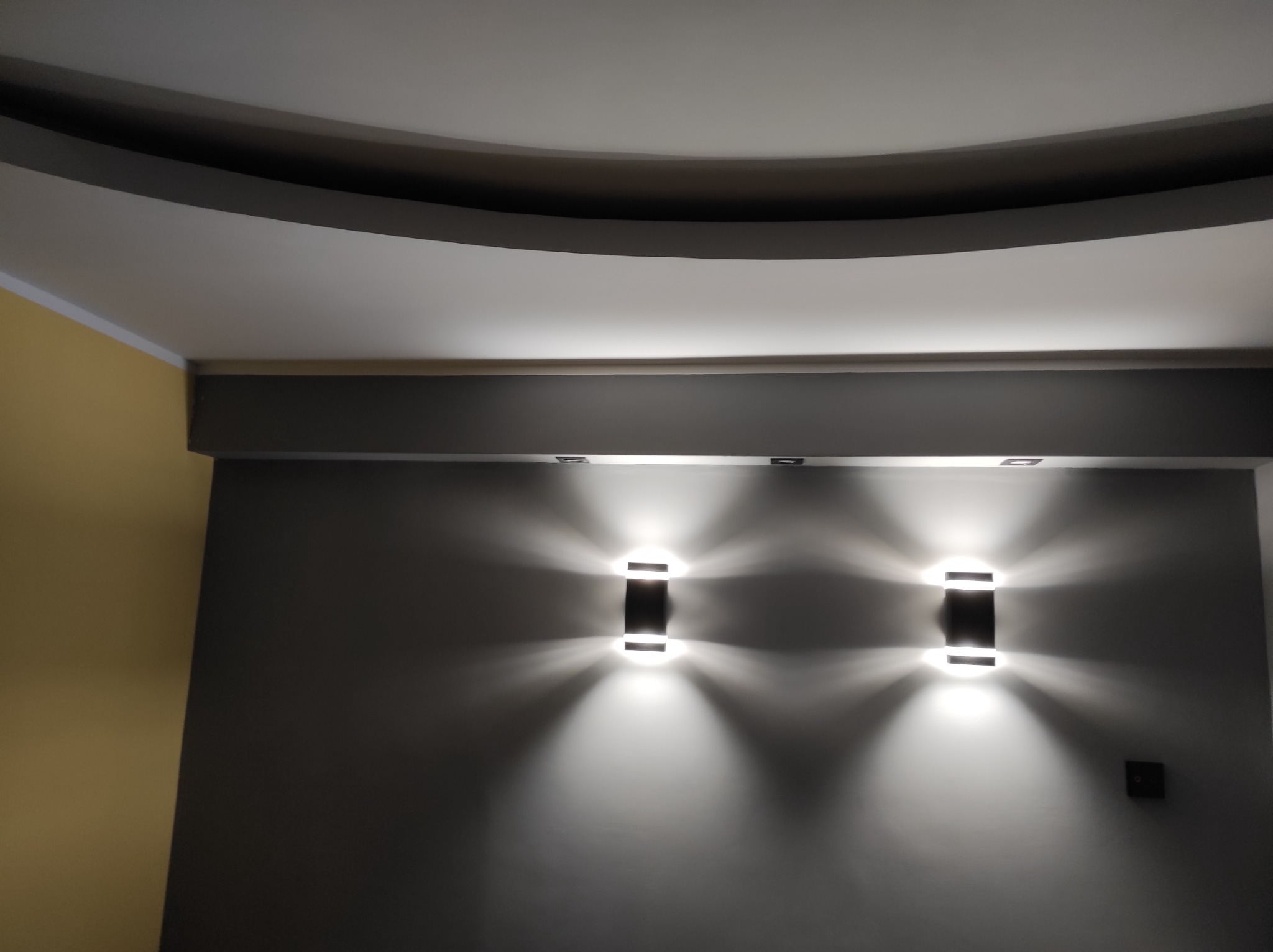 Ściana pomalowana na szaro z dwoma nowoczesnymi kinkietami emitującymi ciepłe światło, widoczny fragment sufitu z dekoracyjnym elementem gipsowym oraz fragment ściany w kolorze musztardowym.