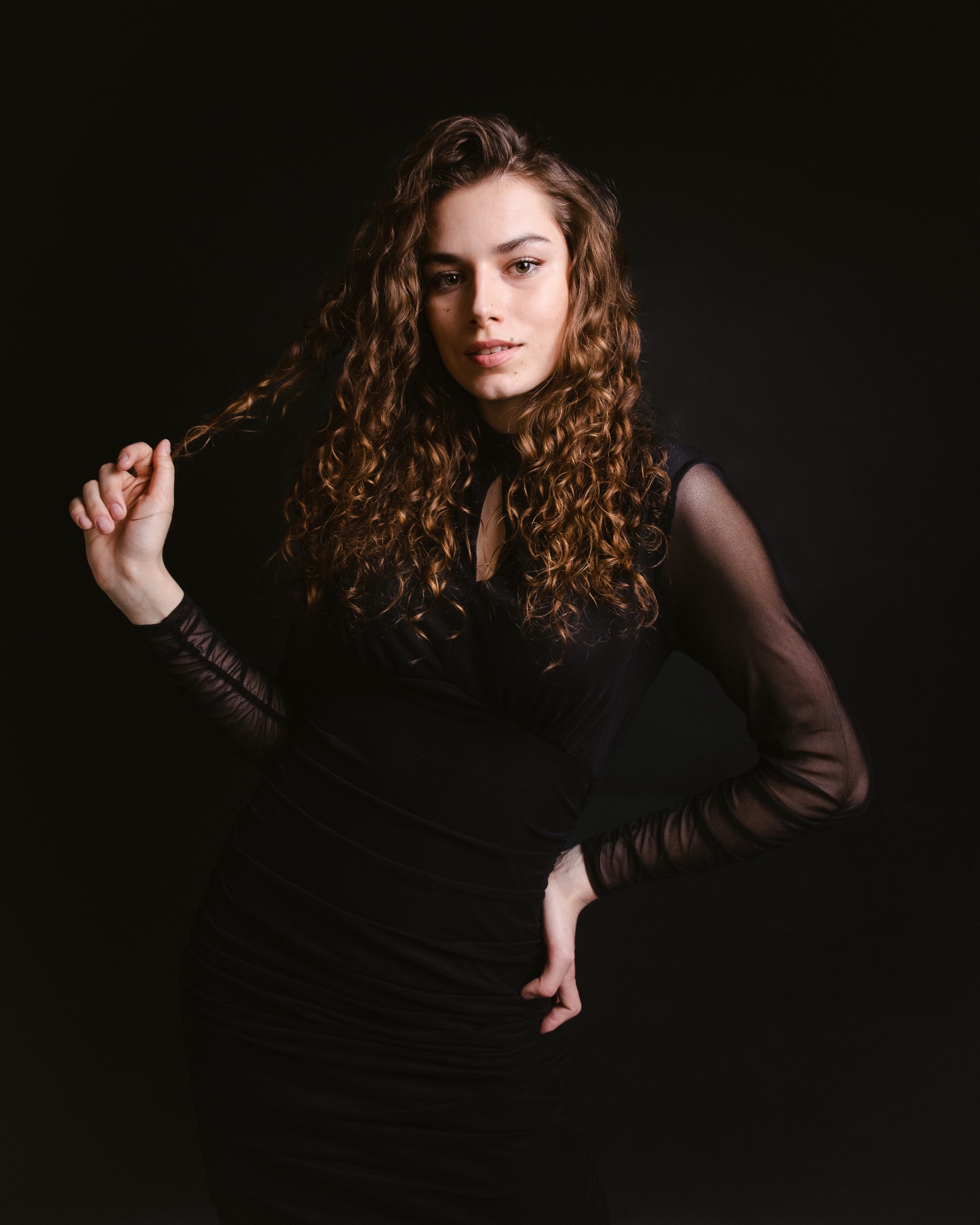 Portret kobiety z kręconymi włosami, ubrana w czarną sukienkę z siateczkowymi rękawami, pozująca na ciemnym tle z dłonią we włosach.