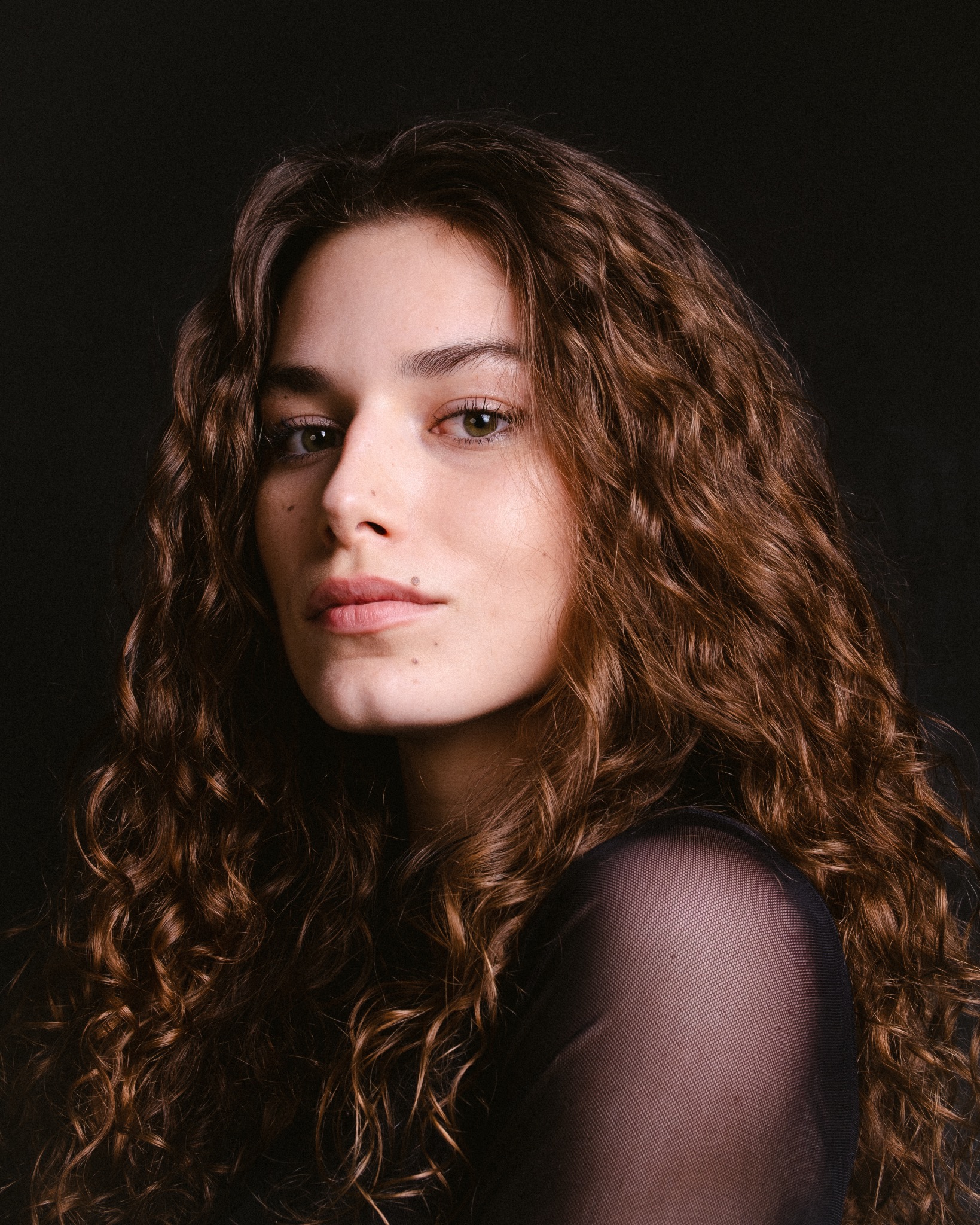 Portret młodej kobiety z długimi, kręconymi włosami na ciemnym tle, z delikatnym makijażem i znamieniem na twarzy.