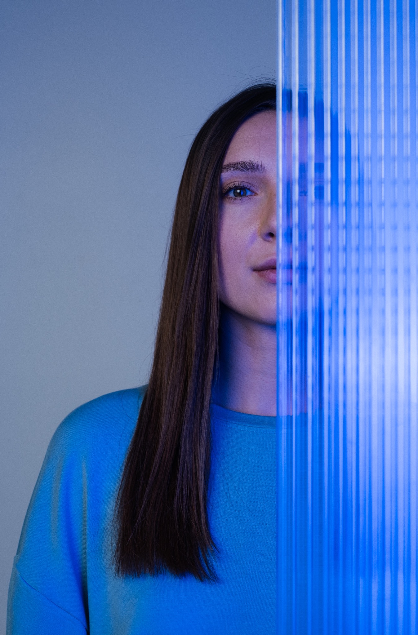Portret kobiety z długimi, prostymi włosami, częściowo zasłoniętej przez niebieskie, pionowe pręty, oświetlonej chłodnym, niebieskim światłem.