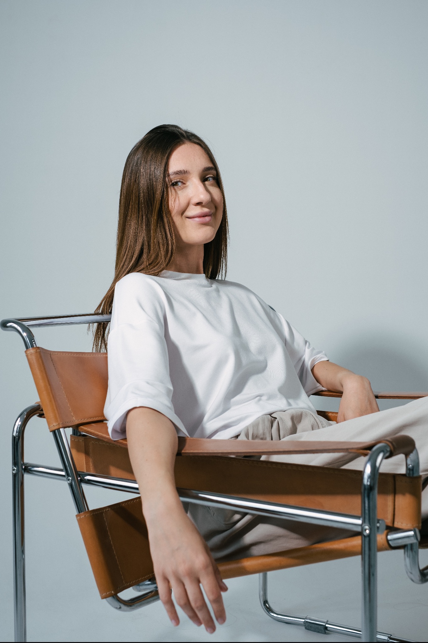 Portret kobiety z długimi brązowymi włosami, ubranej w białą koszulkę i beżowe spodnie, siedzącej na skórzanym krześle Barcelona na tle jasnoszarego studia.