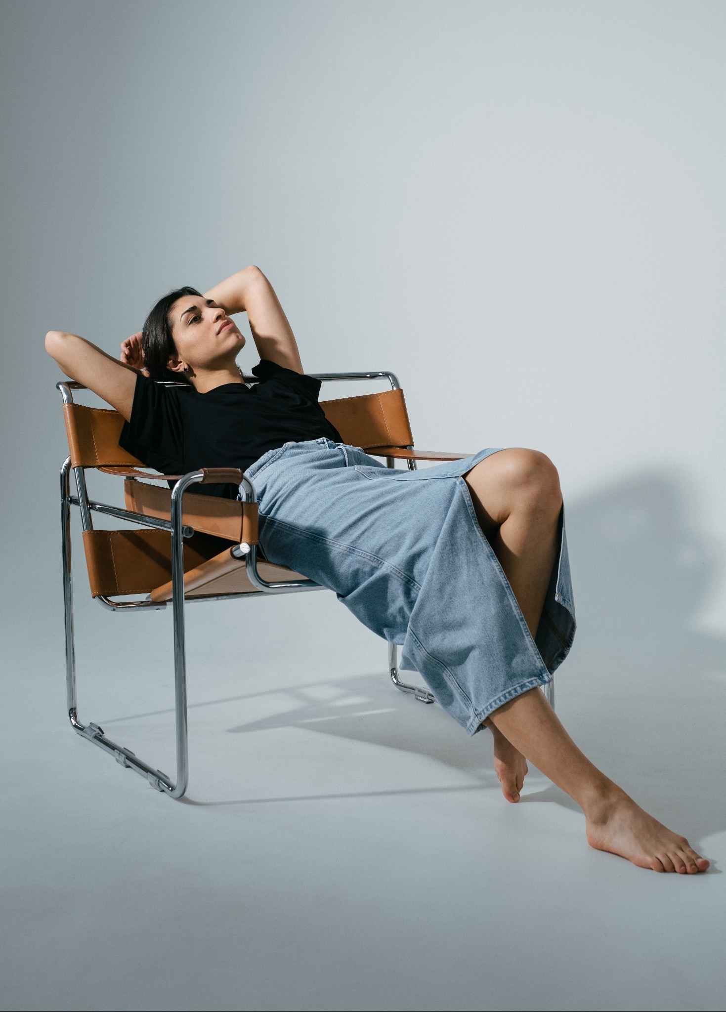 Kobieta w czarnym T-shircie i długiej jeansowej spódnicy relaksuje się na brązowym skórzanym krześle Barcelona na tle jasnoszarego studia.