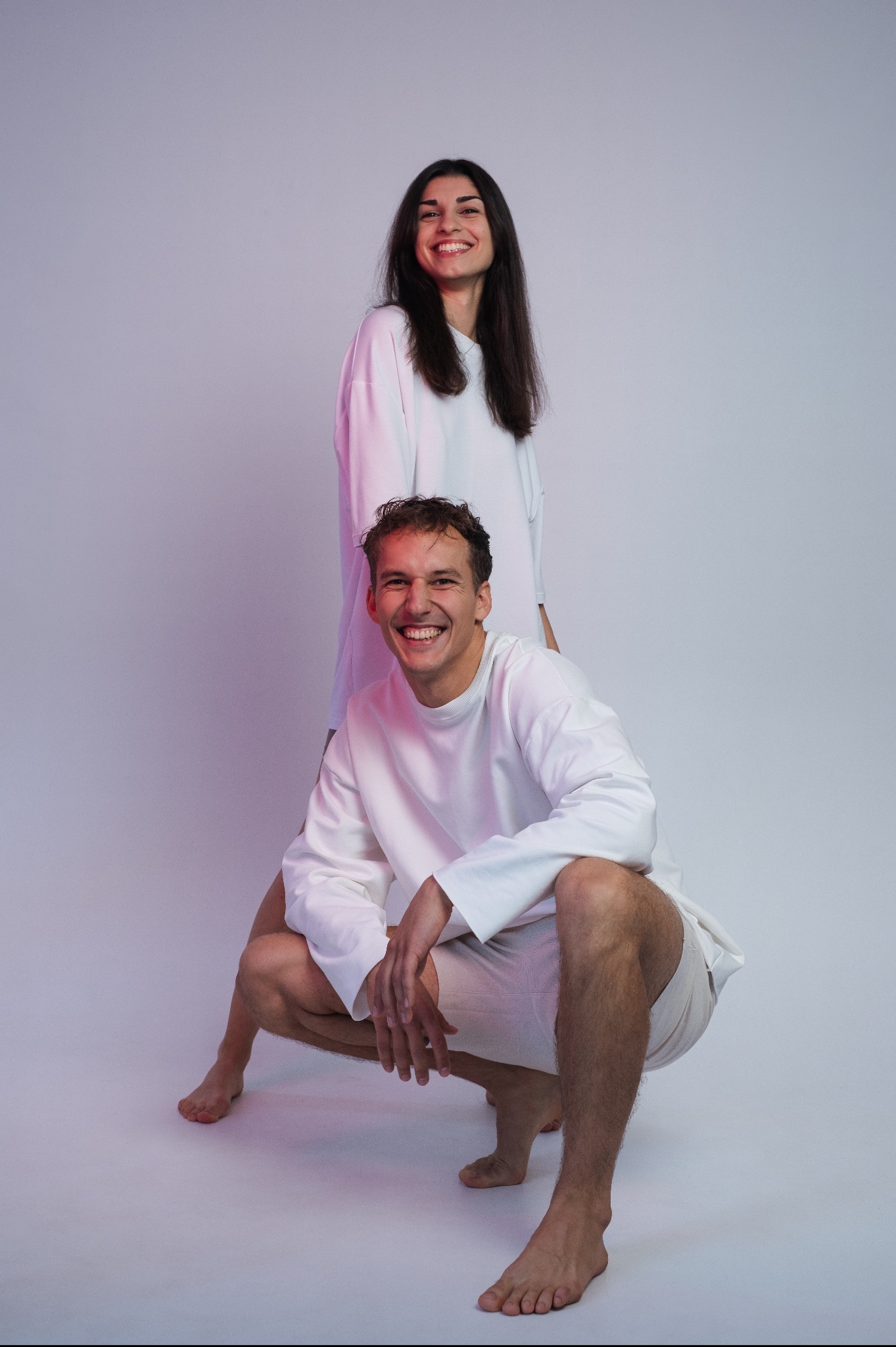 Para w białych ubraniach pozuje w studio w Pruszkowie, mężczyzna kuca, a kobieta stoi za nim, oboje uśmiechnięci, oświetlenie z delikatnym odcieniem różu.