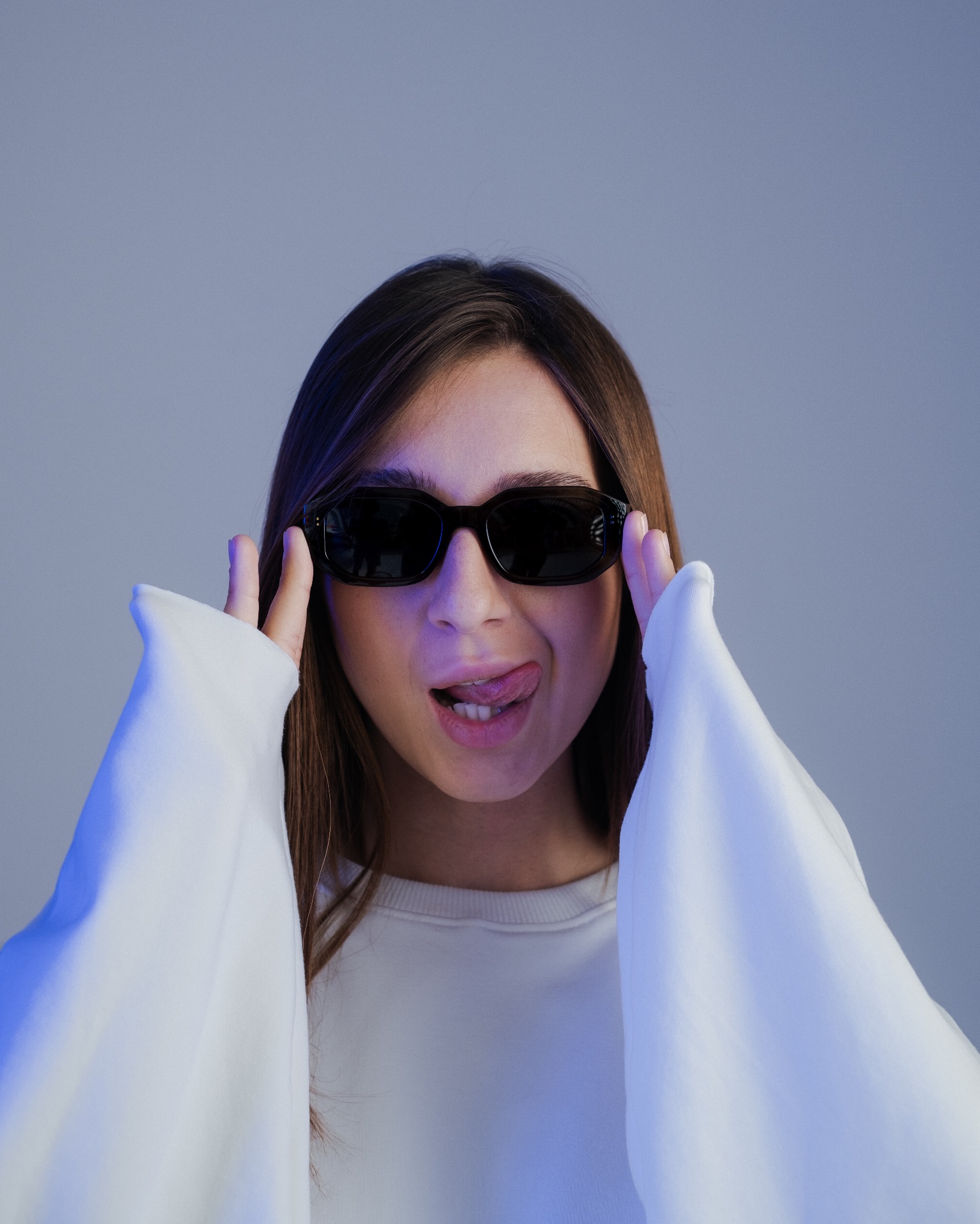 Portret młodej kobiety w czarnych okularach, wystawiającej język, w białej bluzie na tle szarego tła z niebieskim oświetleniem.