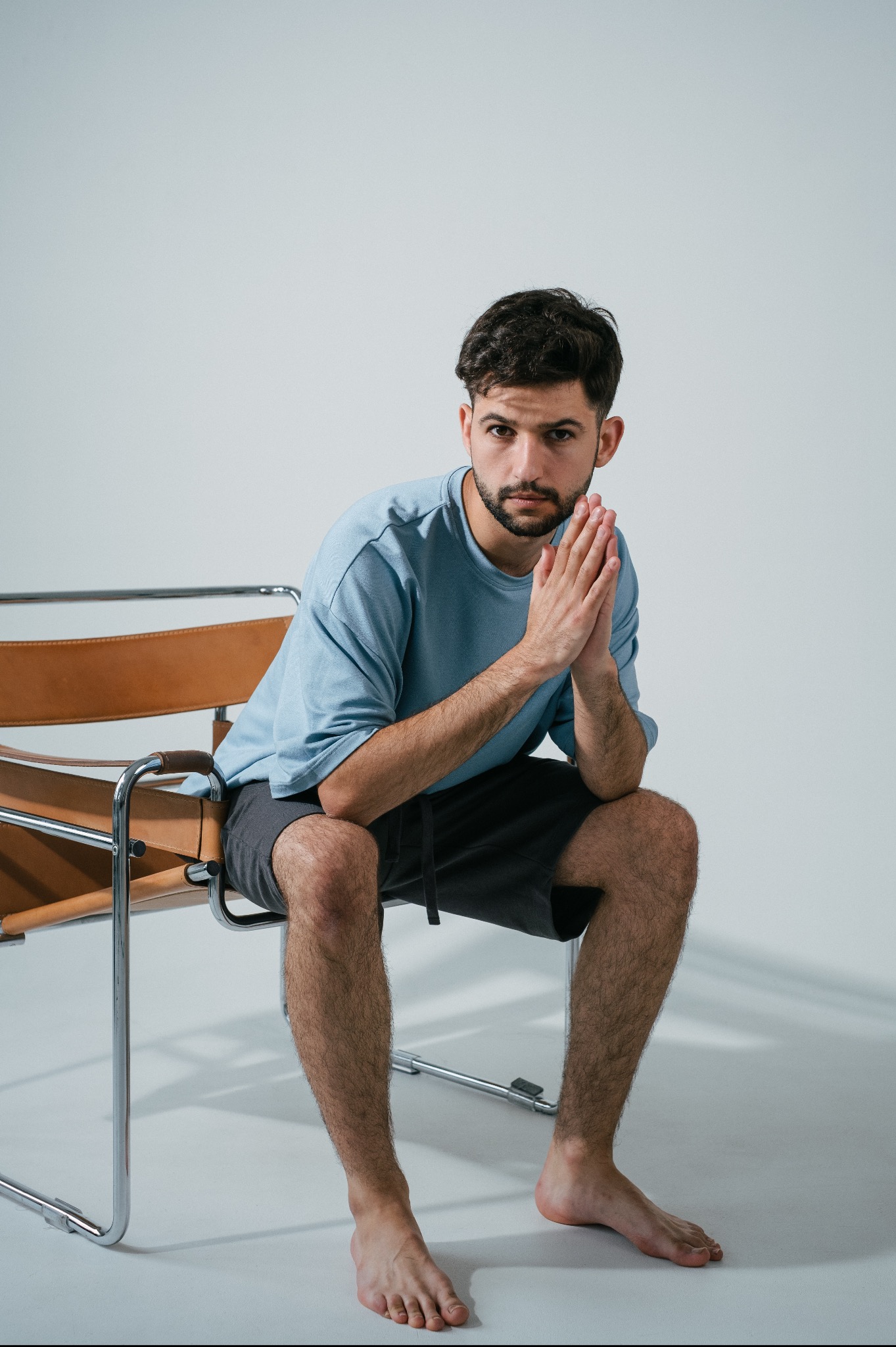 Mężczyzna z brodą siedzi na skórzanym krześle Wassily, z rękami złożonymi przed sobą, ubrany w błękitną koszulkę i czarne spodenki, na jasnym tle.