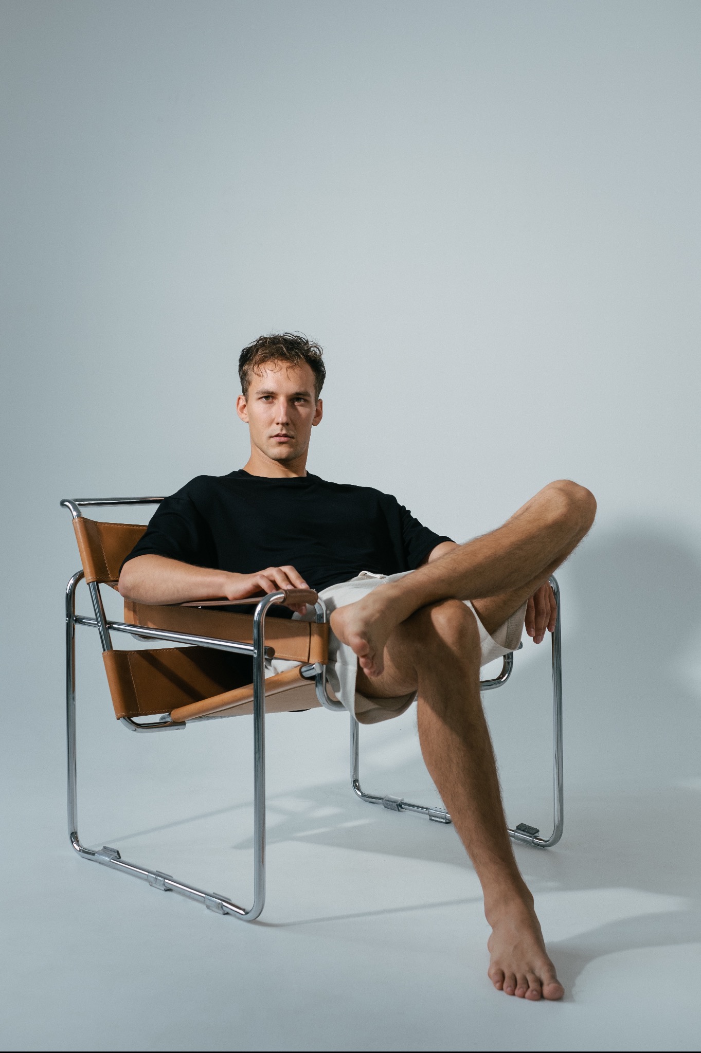Mężczyzna w czarnym T-shircie i jasnych szortach siedzi w skórzanym krześle ze stalową ramą na jasnym tle.