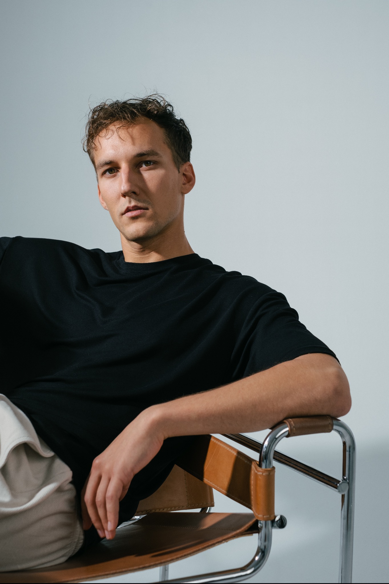 Portret młodego mężczyzny w czarnym T-shircie, siedzącego na skórzanym krześle z metalową ramą, z delikatnym uśmiechem i spojrzeniem w dal.