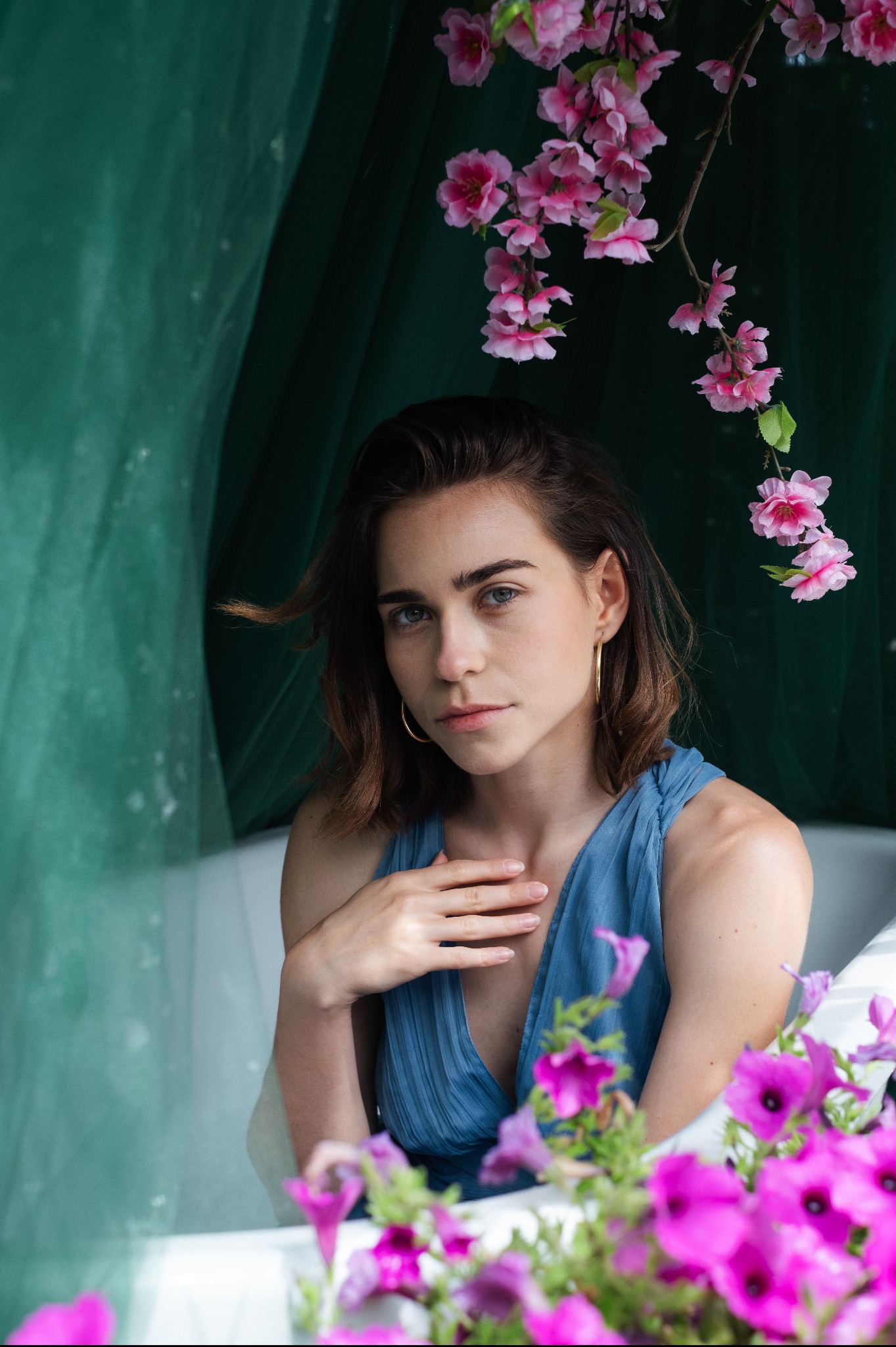 Portret kobiety w niebieskiej sukience, otoczonej kwiatami i zielonym materiałem, z delikatnym makijażem i złotymi kolczykami.