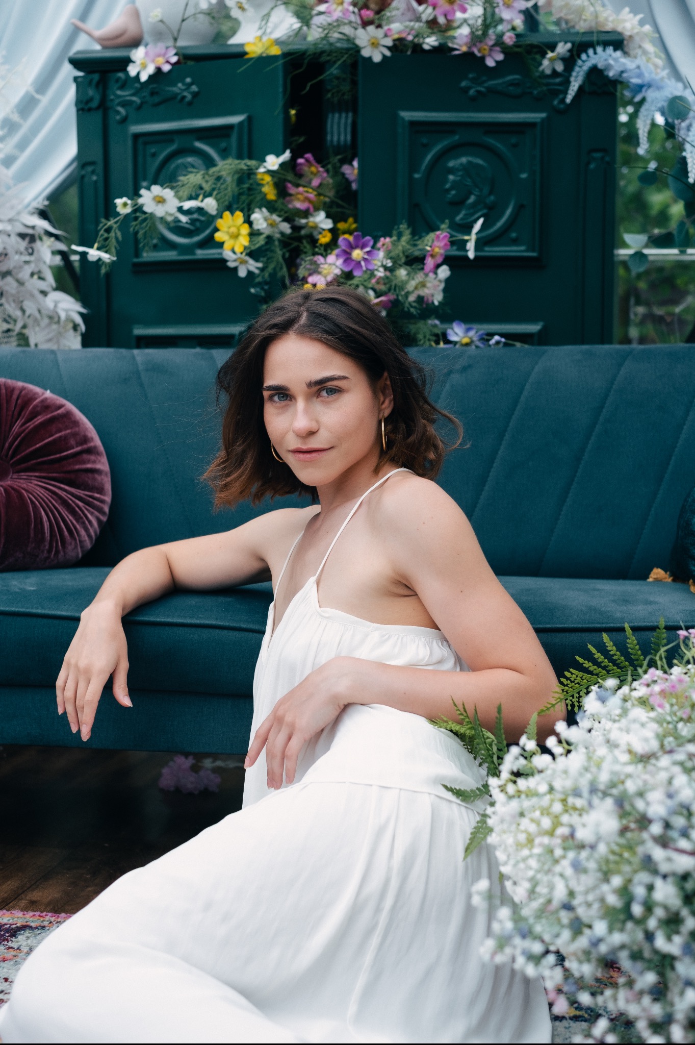 Portret kobiety w białej sukience, siedzącej na podłodze przed turkusową kanapą, z kwiatami i ozdobnymi drzwiami w tle.