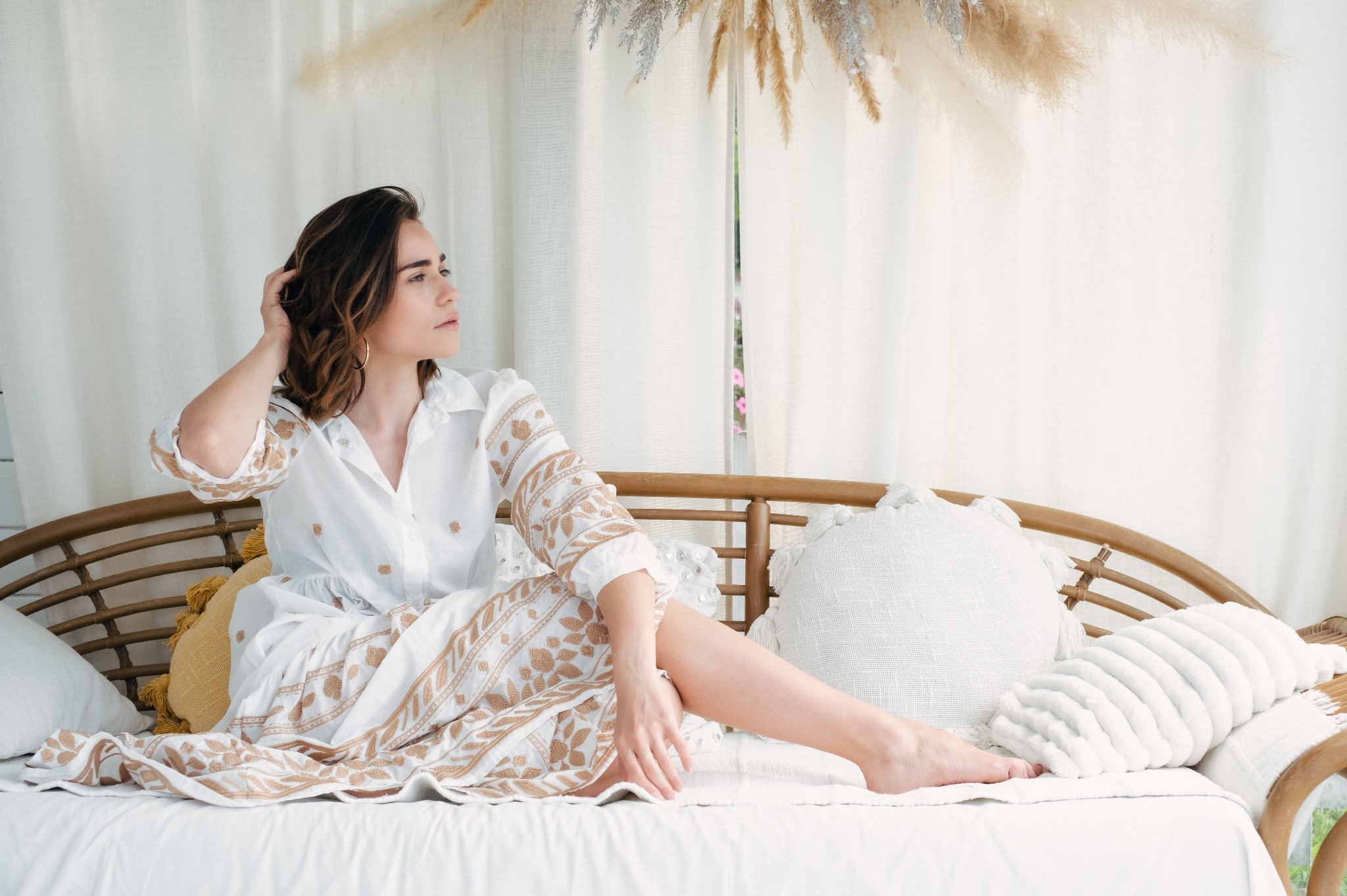 Kobieta w białej, haftowanej sukience siedzi na rattanowej sofie z poduszkami, odwrócona w bok, z ręką we włosach, na tle białych zasłon i dekoracji z suszonych traw.