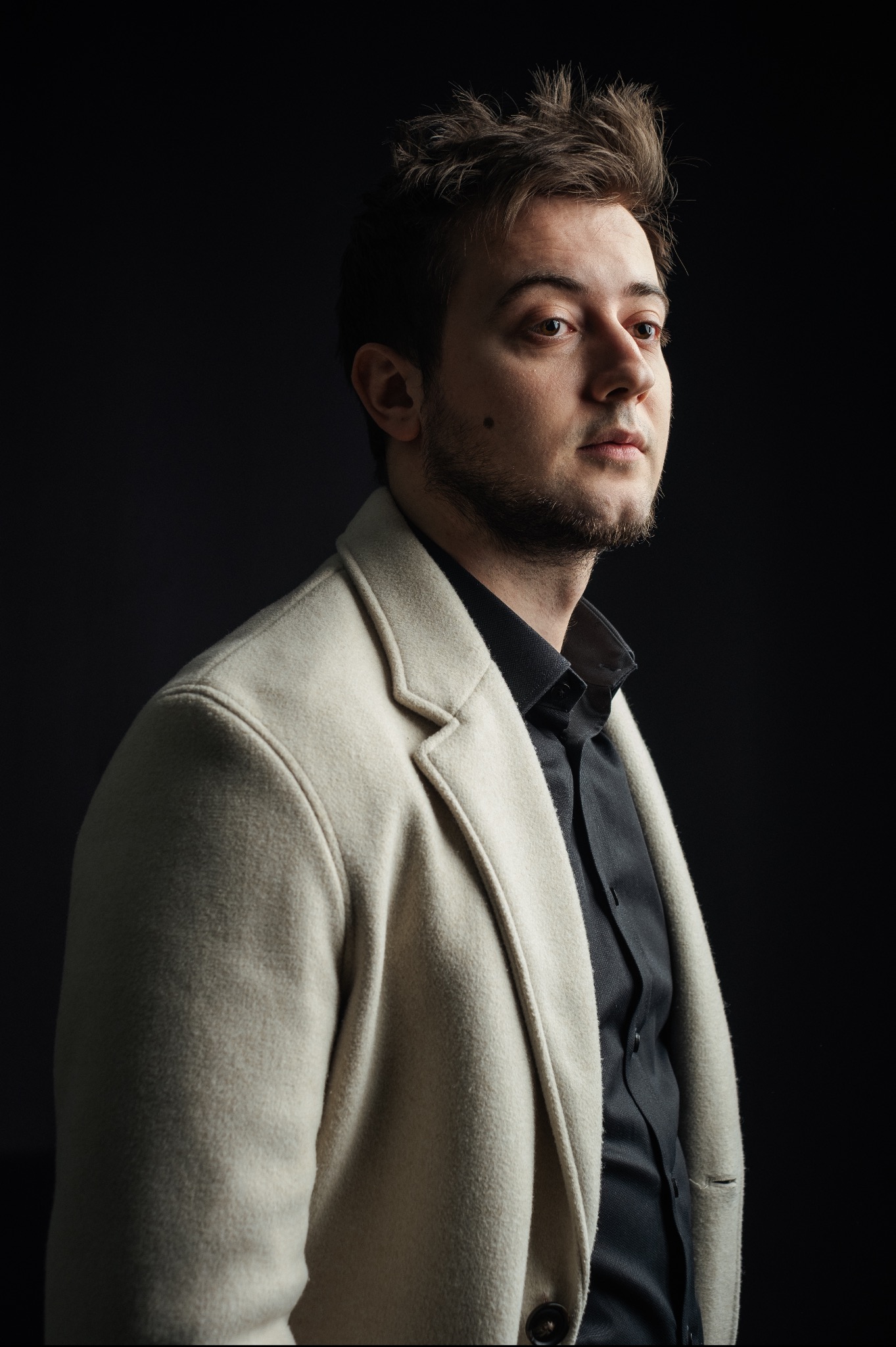 Portret mężczyzny w jasnym płaszczu i czarnej koszuli na ciemnym tle, oświetlony z boku, z lekkim zarostem i charakterystycznym pieprzykiem na policzku.