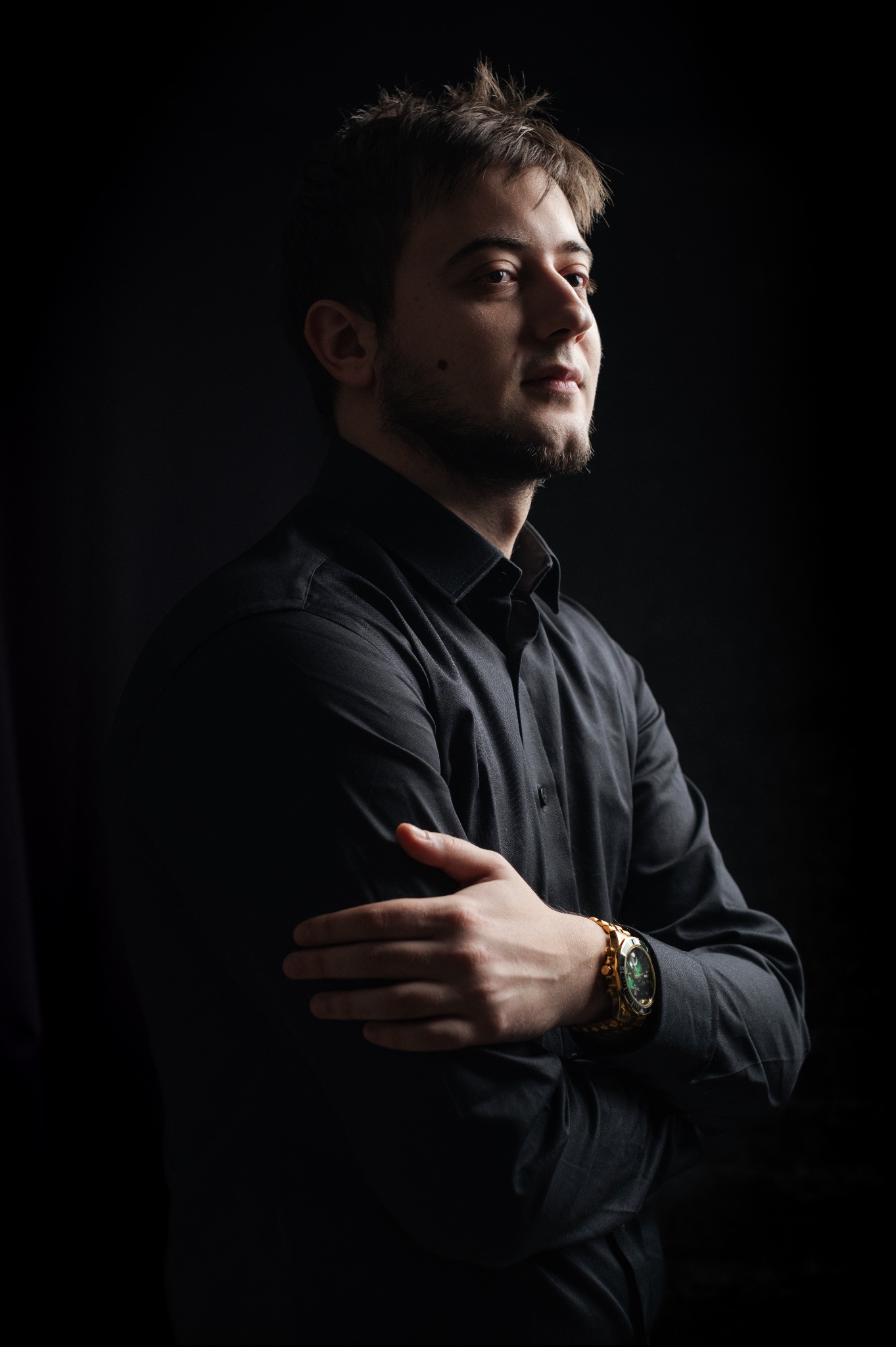 Mężczyzna w czarnej koszuli, z rękami skrzyżowanymi na piersiach, patrzący w prawo, z widocznym złotym zegarkiem na nadgarstku, na ciemnym tle.