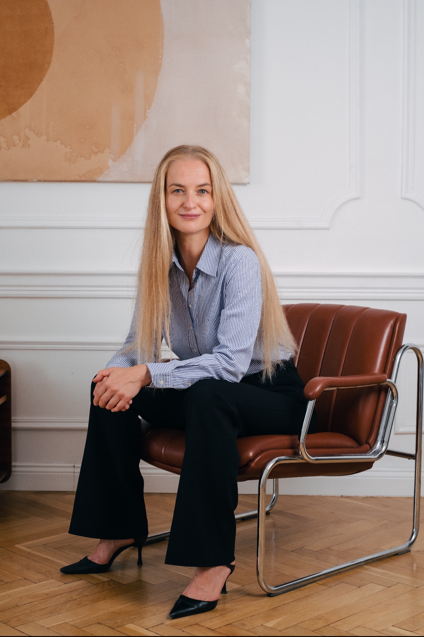 Portret kobiety z długimi blond włosami, siedzącej na brązowym skórzanym krześle w paski, w niebieskiej koszuli w paski i czarnych spodniach, na drewnianej podłodze, z abstrakcyjnym obrazem w tle.