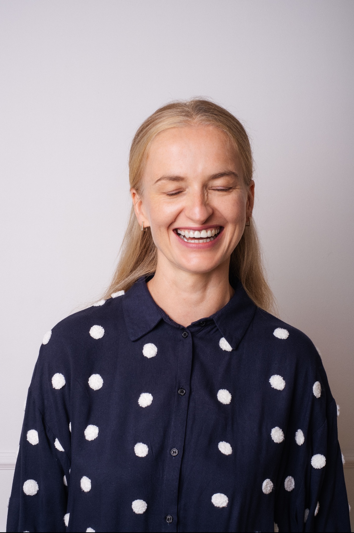 Portret roześmianej blondynki z zamkniętymi oczami, ubrana w granatową koszulę w białe kropki.