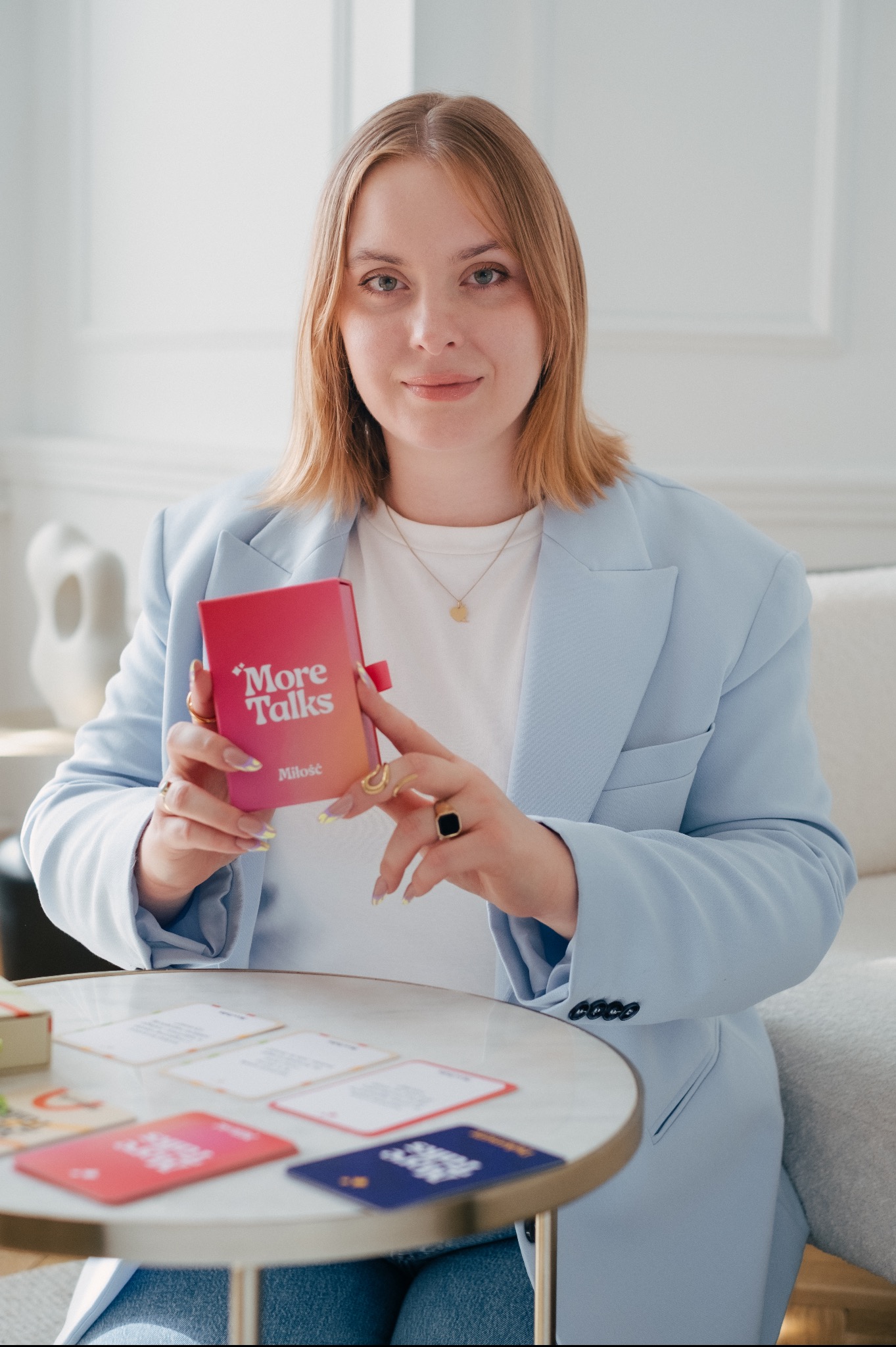 Kobieta w błękitnym żakiecie prezentuje grę karcianą 'More Talks Miłość' siedząc przy stoliku z rozłożonymi kartami.