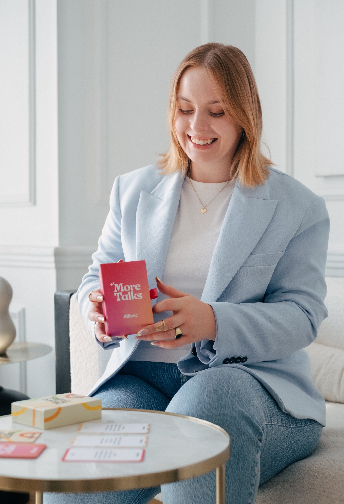 Uśmiechnięta blondynka w błękitnej marynarce siedzi na kanapie i trzyma w dłoniach pudełko gry karcianej 'More Talks Miłość', na stoliku leżą karty z pytaniami.