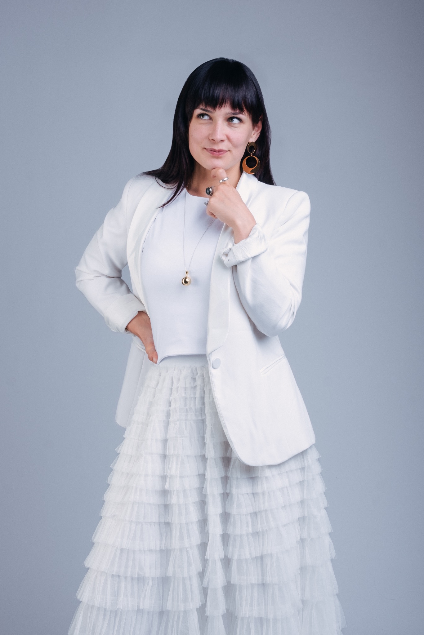 Kobieta w białym komplecie, z ręką pod brodą, spogląda w lewo, ubrana w białą marynarkę, bluzkę i tiulową spódnicę, z wiszącymi kolczykami i naszyjnikiem z kulką.
