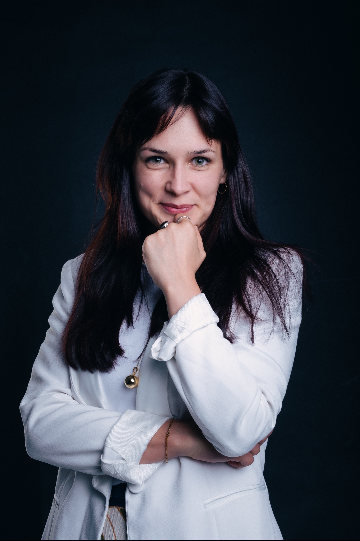 Portret uśmiechniętej kobiety w białej marynarce, z ręką pod brodą, na ciemnym tle, z widoczną biżuterią: pierścionkami, kolczykami i naszyjnikiem z kulką.