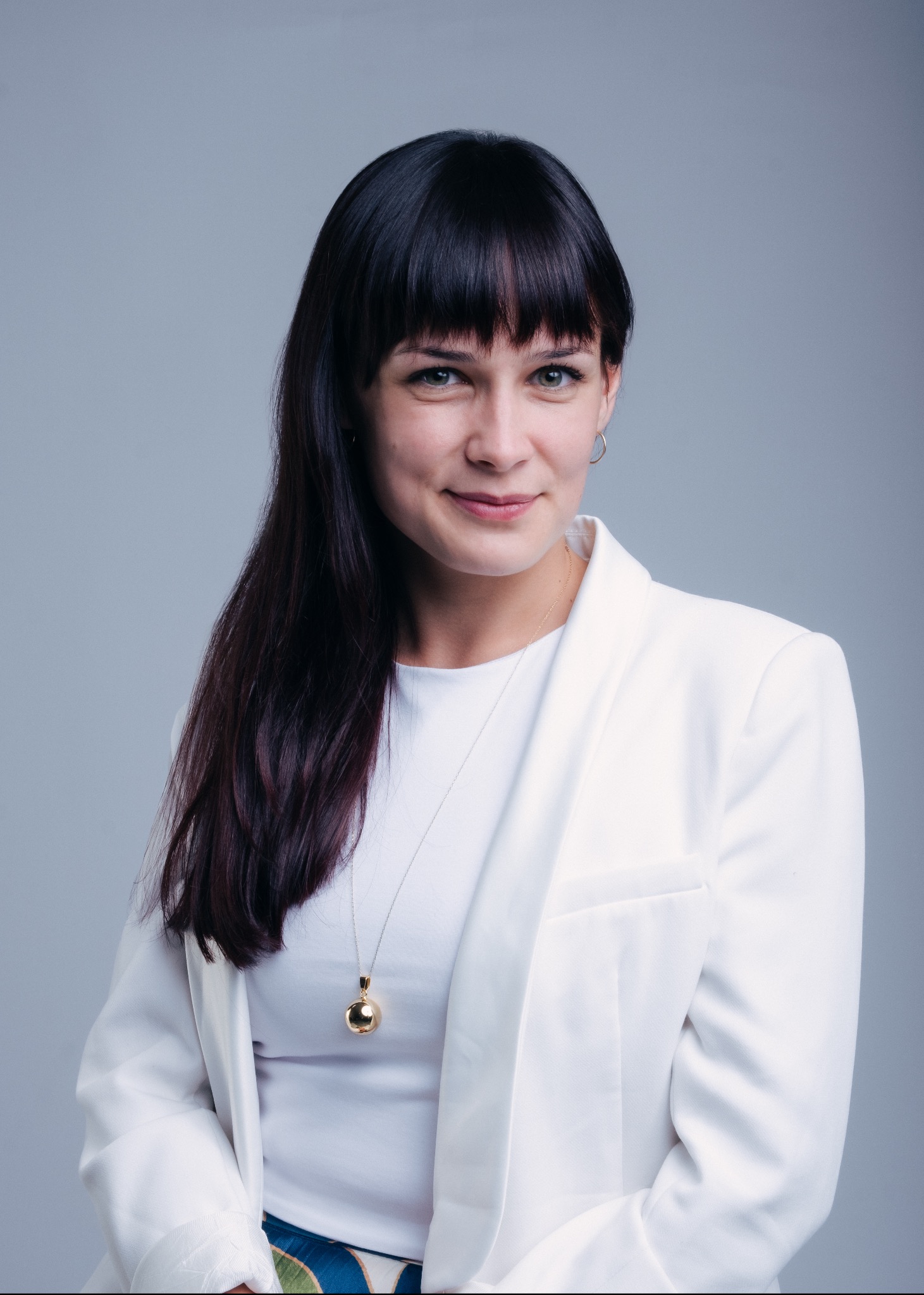 Portret uśmiechniętej kobiety z ciemnymi włosami i grzywką, ubrana w białą marynarkę i bluzkę, z złotym naszyjnikiem na szarym tle.