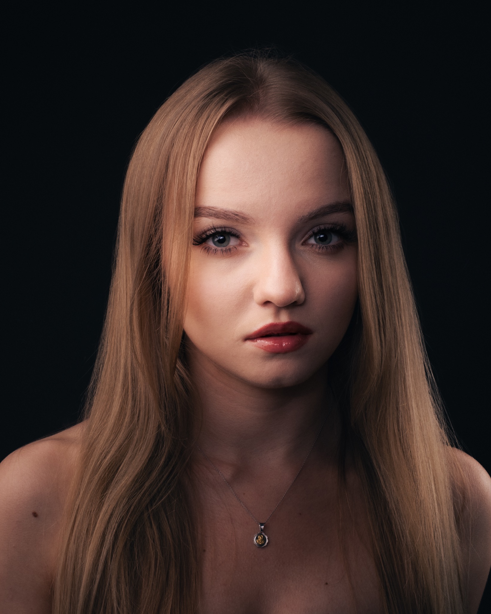 Portret młodej kobiety z długimi, prostymi blond włosami, w srebrnym naszyjniku z żółtym kamieniem, na ciemnym tle.