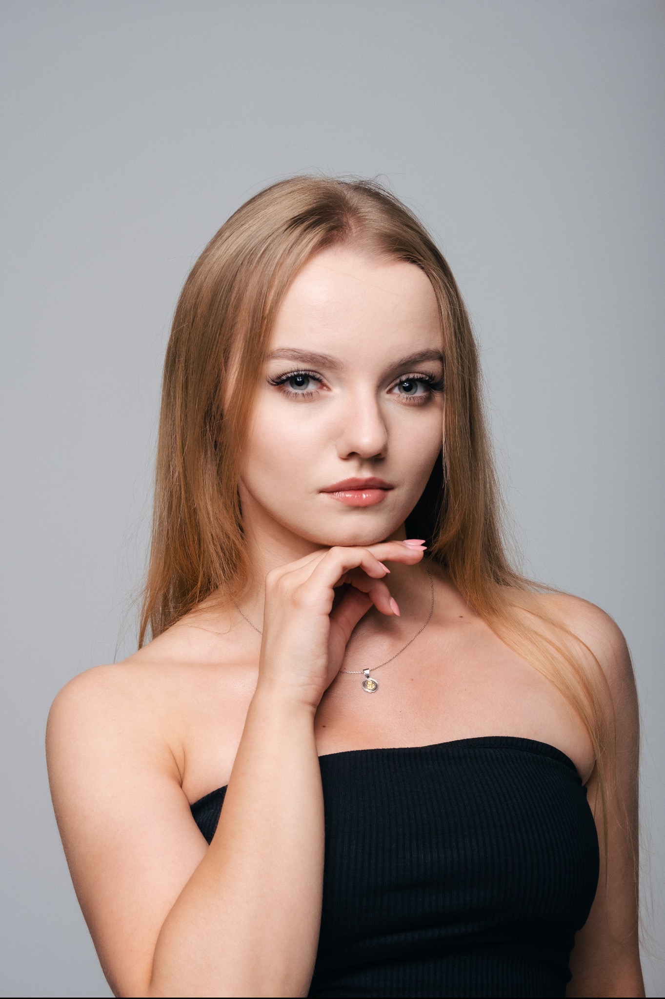Portret młodej kobiety z blond włosami, w czarnym topie, z ręką pod brodą i delikatnym srebrnym naszyjnikiem na szarym tle.