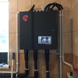 Euros Energy Sp. z o.o JN - Czarna obudowa urządzenia Eurocontrol z podłączonymi rurami, zamontowana na kafelkowanej ścianie w pomieszczeniu technicznym, widoczny ekran z temperaturą 46 stopni Celsjusza, obok pionowa biała...