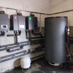 Euros Energy Sp. z o.o JN - Trzy szare urządzenia z różowymi i zielonymi logotypami na ścianie, połączone czarnymi, izolowanymi rurami, obok duży, czarny zbiornik akumulacyjny i mniejszy, biały zbiornik.